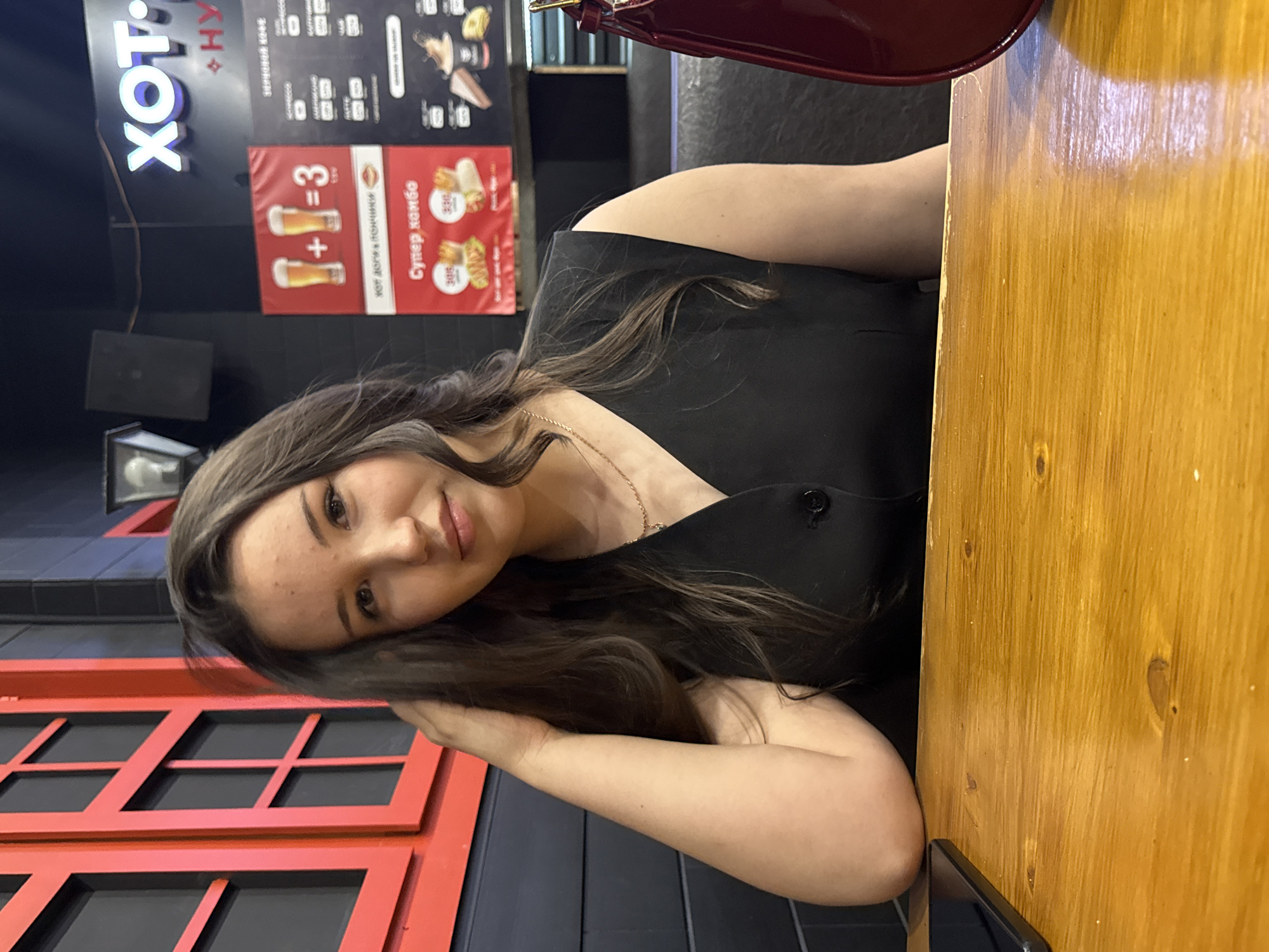 A young woman with long, dark hair is seated at a wooden table in what appears to be a fast-food establishment in Perm, Russia. She is wearing a black sleeveless top. Her right arm is bent, with her hand resting on her head, and her left arm is resting on the table. In the background, there is a red window frame with black panes, a dark wall, and a menu board with neon signs advertising food and drinks. The menu board displays images of beer and food items, with prices indicated in Russian currency. A dark object, possibly a phone, is visible on the table to the left. To the right of the woman, a red handbag is partially visible. The lighting suggests an indoor environment, possibly artificial.