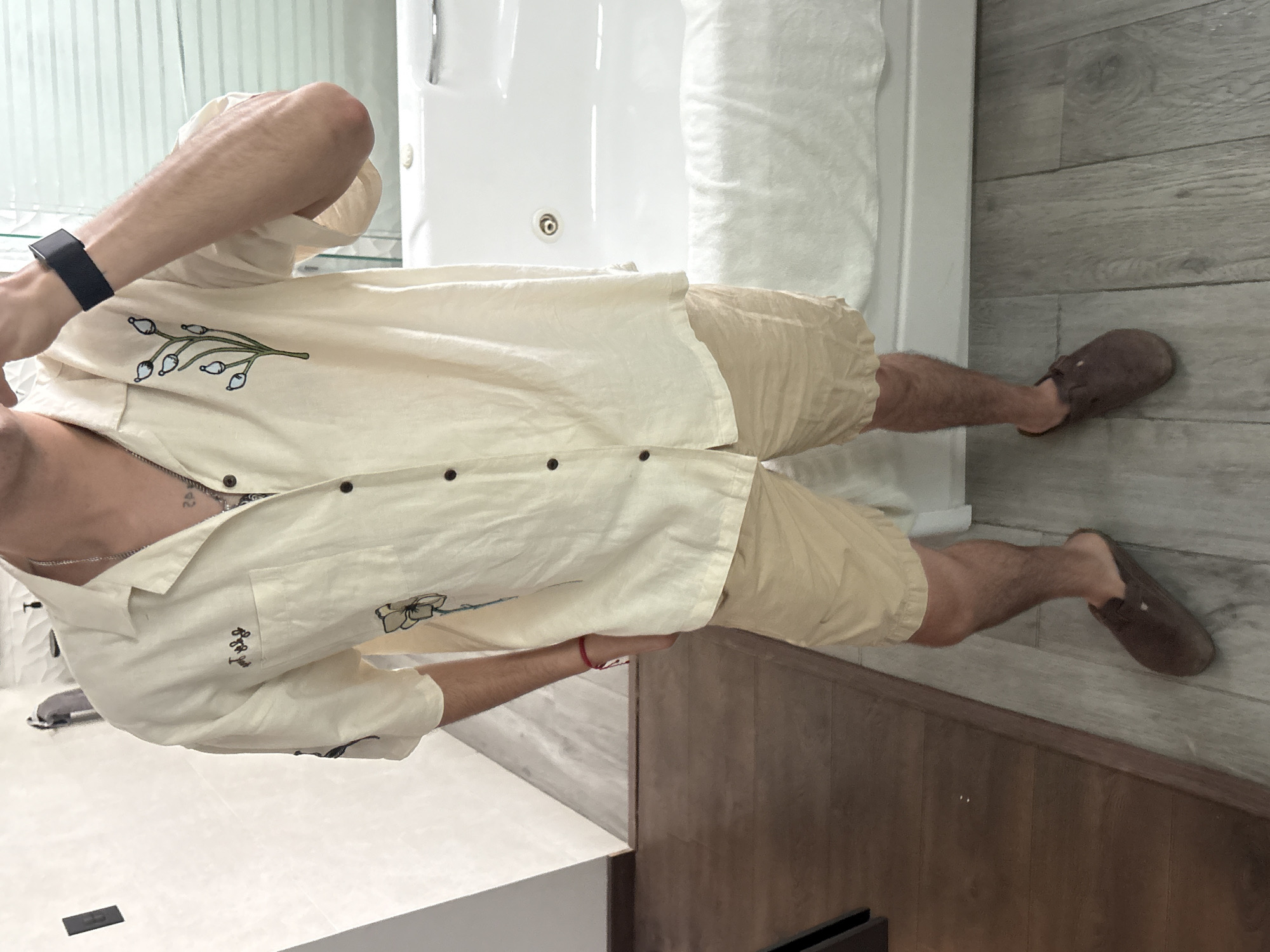 A person wearing a light-colored button-down shirt with floral designs, tan shorts, and brown slippers stands in a bathroom. The bathroom features a white jacuzzi tub with a rolled white towel on it, grey wood-look flooring, and dark wood cabinetry. The location is not verifiable as San Bernardino Tlaxcalancingo, Mexico.