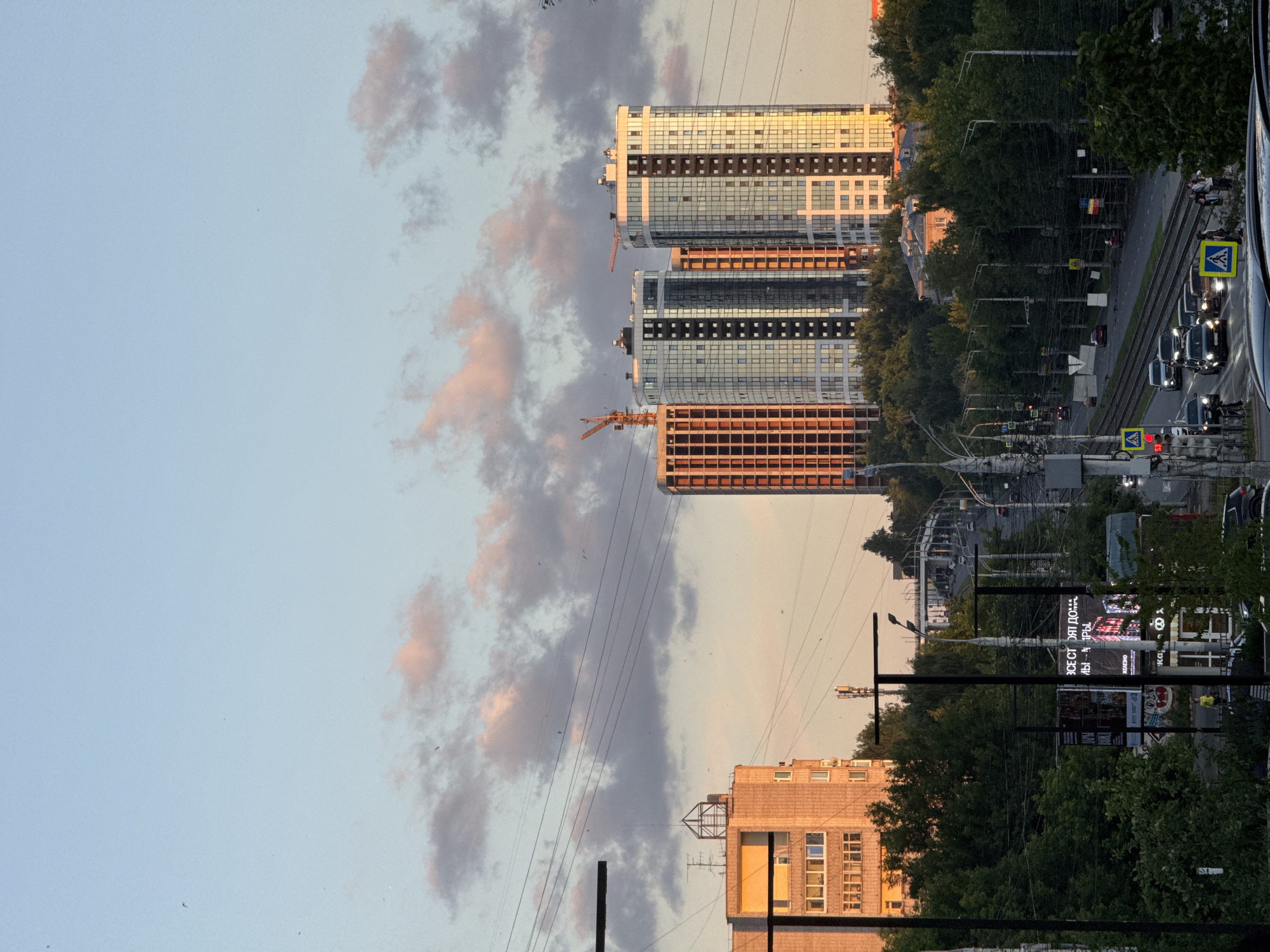 This image captures a city scene in Izhevsk, Russia, likely during the late afternoon or early evening, as indicated by the warm, golden light illuminating the buildings and the soft hues in the sky. The perspective is from a slightly elevated viewpoint, looking down a street lined with trees and passing traffic.
Dominating the background are several modern high-rise buildings, some of which appear to be under construction, evidenced by exposed frameworks and cranes. The completed structures feature reflective glass facades that catch the light, creating a shimmering effect. In the mid-ground, older, brick buildings stand, and a network of power lines crisscrosses the frame, typical of urban landscapes.
The street below is active with vehicles, including cars and possibly trams, given the presence of tracks. Pedestrians are visible on the sidewalks and near a crosswalk, some engaged in conversation. The surrounding environment is lush with green trees, suggesting a temperate climate. The sky is a soft blue, interspersed with scattered clouds tinged with pink and orange, further confirming the time of day. Signage in Russian is visible on billboards and poles.
