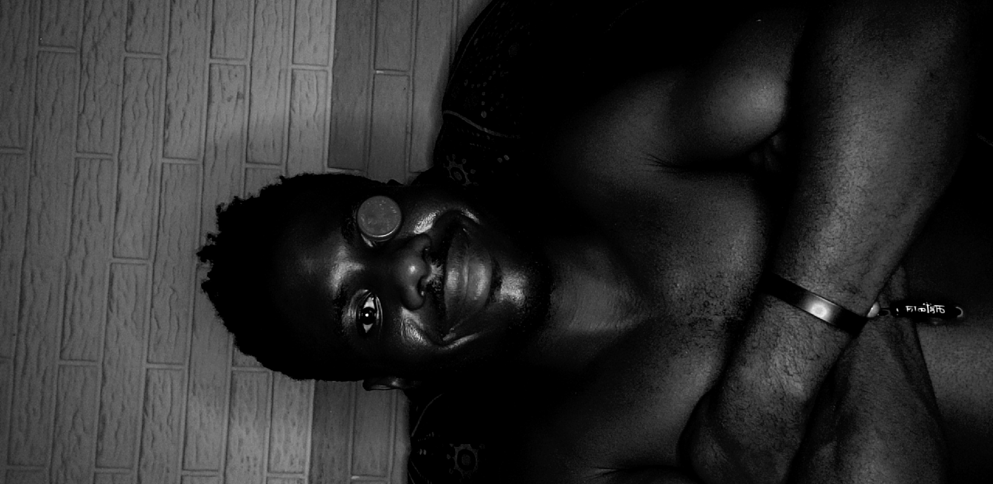 A black and white, vertically oriented photograph captures a person lying down, engaging with the camera with a wide smile. The subject is a dark-skinned individual with short, curly hair and some facial stubble, illuminated by strong, directional light. A circular metallic object, possibly a coin, is balanced on their forehead.
The lighting creates dramatic highlights on the person's forehead, nose, cheekbones, lips, and left shoulder, accentuating their features and muscular build, while casting much of the rest of their body into deep shadow. Their left eye is bright and open, looking directly at the viewer, conveying a sense of playfulness or happiness.
The background on the left features a light-toned wall with a textured, rectangular brick-like or tiled pattern. Beneath the subject's head and shoulder, a dark fabric or cushion with a subtle, intricate pattern is visible. On the person's right wrist, a dark, thick wristband is worn. White characters in a non-Latin script, along with an exclamation mark and a closing parenthesis, are visible on the wristband.
The scene appears to be an indoor setting in Narayi, Nigeria, with the activity focused on the subject's expressive face and the balancing act of the coin.