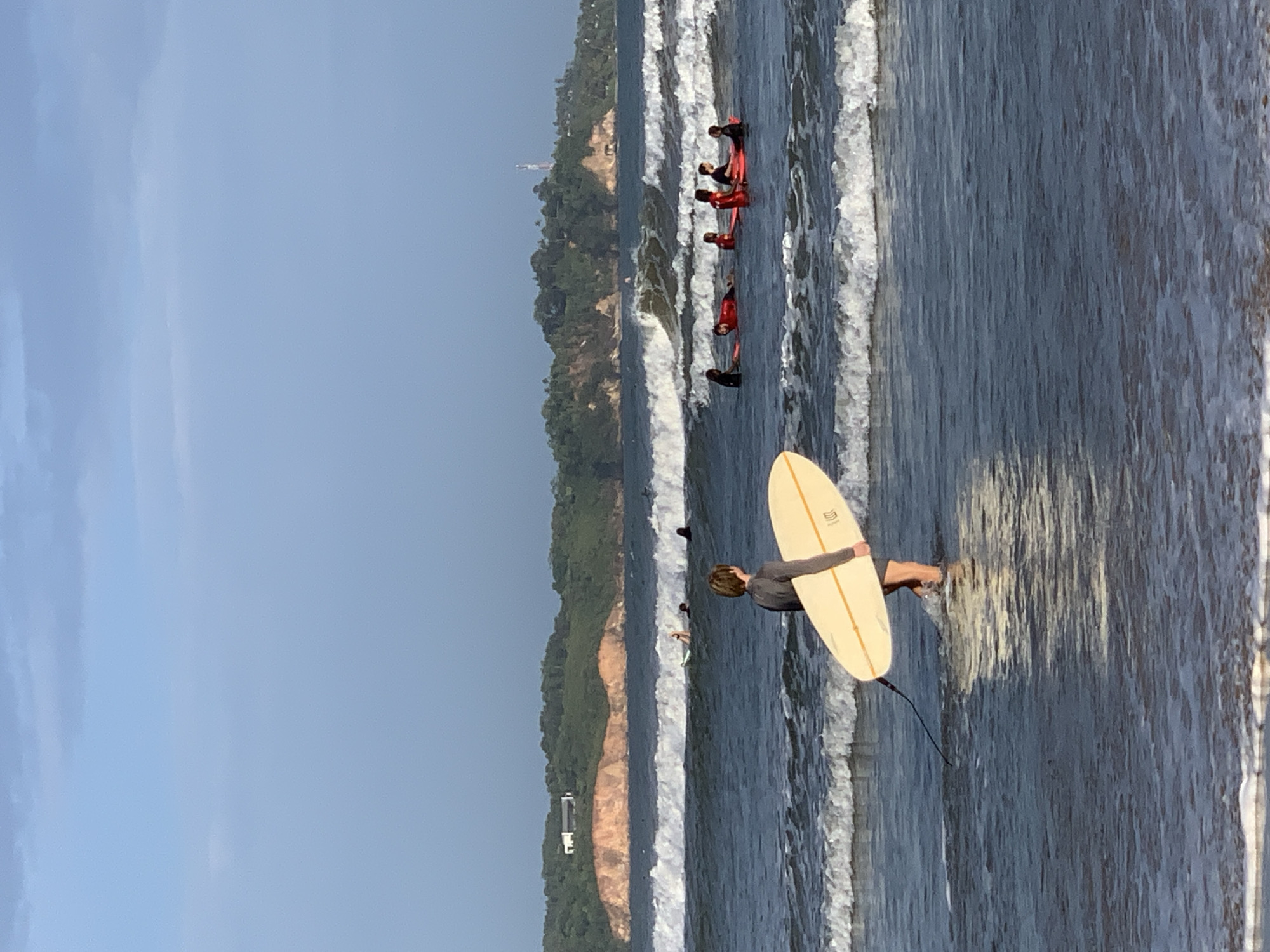 A medium shot depicts a person holding a surfboard while standing in the ocean at Weligama, Sri Lanka. The person, viewed from the back, stands waist-deep in the water, facing the oncoming waves. They are holding a light yellow surfboard horizontally in front of them. Several surfers, wearing red wetsuits, are paddling on their surfboards further out in the ocean. In the background, a coastline is visible with buildings and vegetation, likely located in Weligama. The sky is clear and blue.