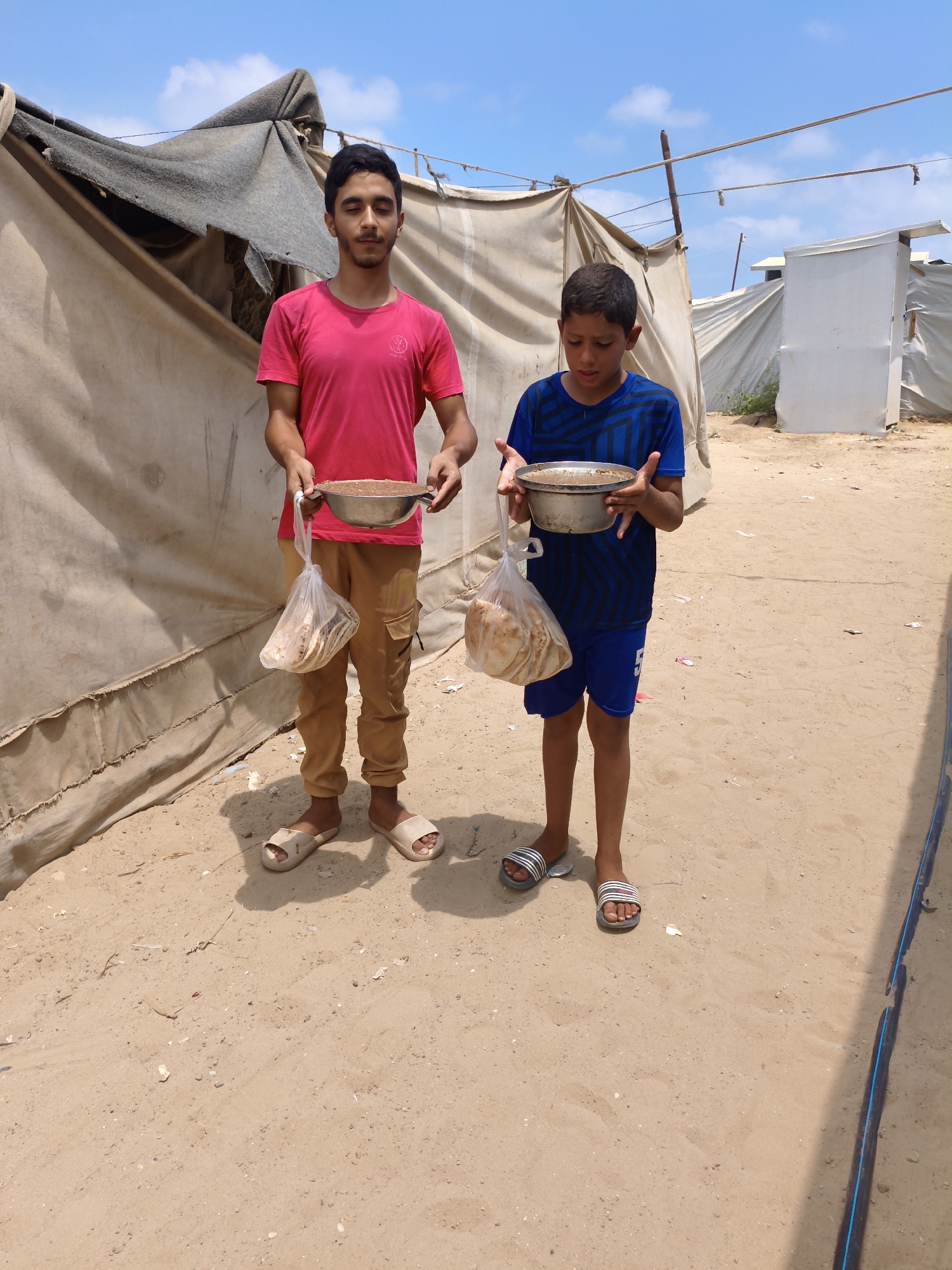 In a sandy, outdoor area, two young males stand holding metal bowls filled with food and plastic bags containing bread. The male on the left wears a bright pink t-shirt and tan cargo pants, while the male on the right is dressed in a blue and black patterned t-shirt and blue shorts. They are positioned in front of a tent-like structure, with more tents and buildings visible in the background under a bright blue sky with scattered clouds. The ground is covered in sand, with some debris scattered around.