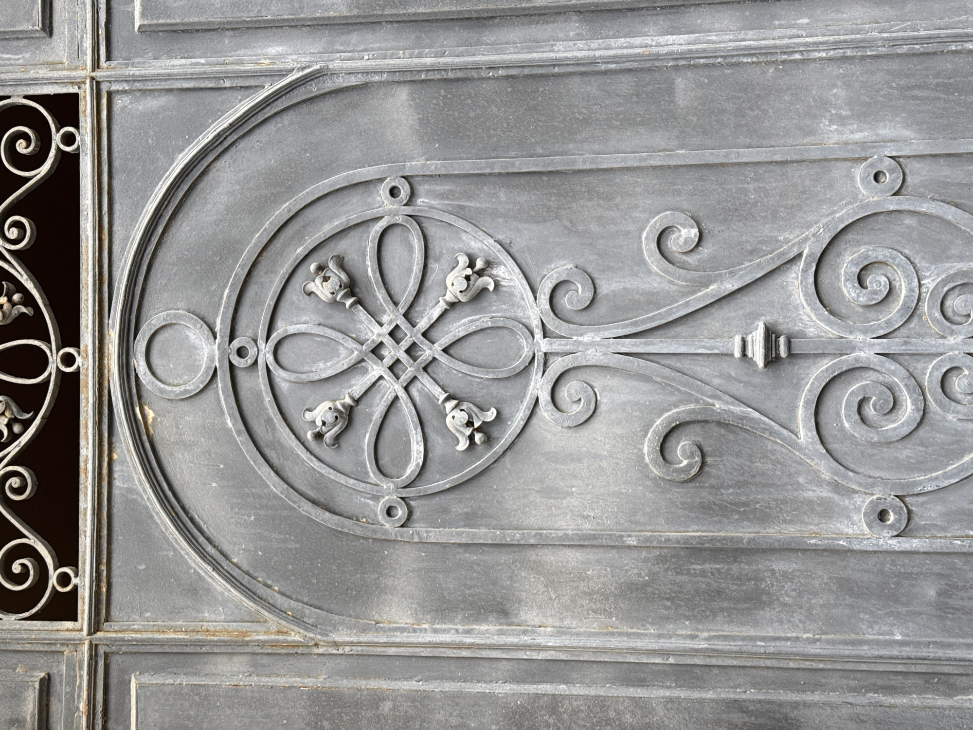 The image shows a close-up of an ornate metal gate, likely part of an entrance to a building or courtyard in Saint Petersburg, Russia. The gate is made of dark grey metal, with visible signs of age and weathering, including some rust spots and a textured surface. The central decorative element is a complex geometric pattern featuring interwoven circular and oval shapes, culminating in stylized floral motifs that resemble tulips or buds at the ends of radiating lines. This central design is enclosed within a larger arched frame, also made of metal. Below the central medallion, scrollwork and curlicue elements further adorn the gate. The top section of the image shows a glimpse of a decorative metal grille, hinting at the upper part of the gate or an adjoining fence. The overall impression is one of historical architecture and craftsmanship. There are no people or actions depicted in the image, only the static structure of the gate.