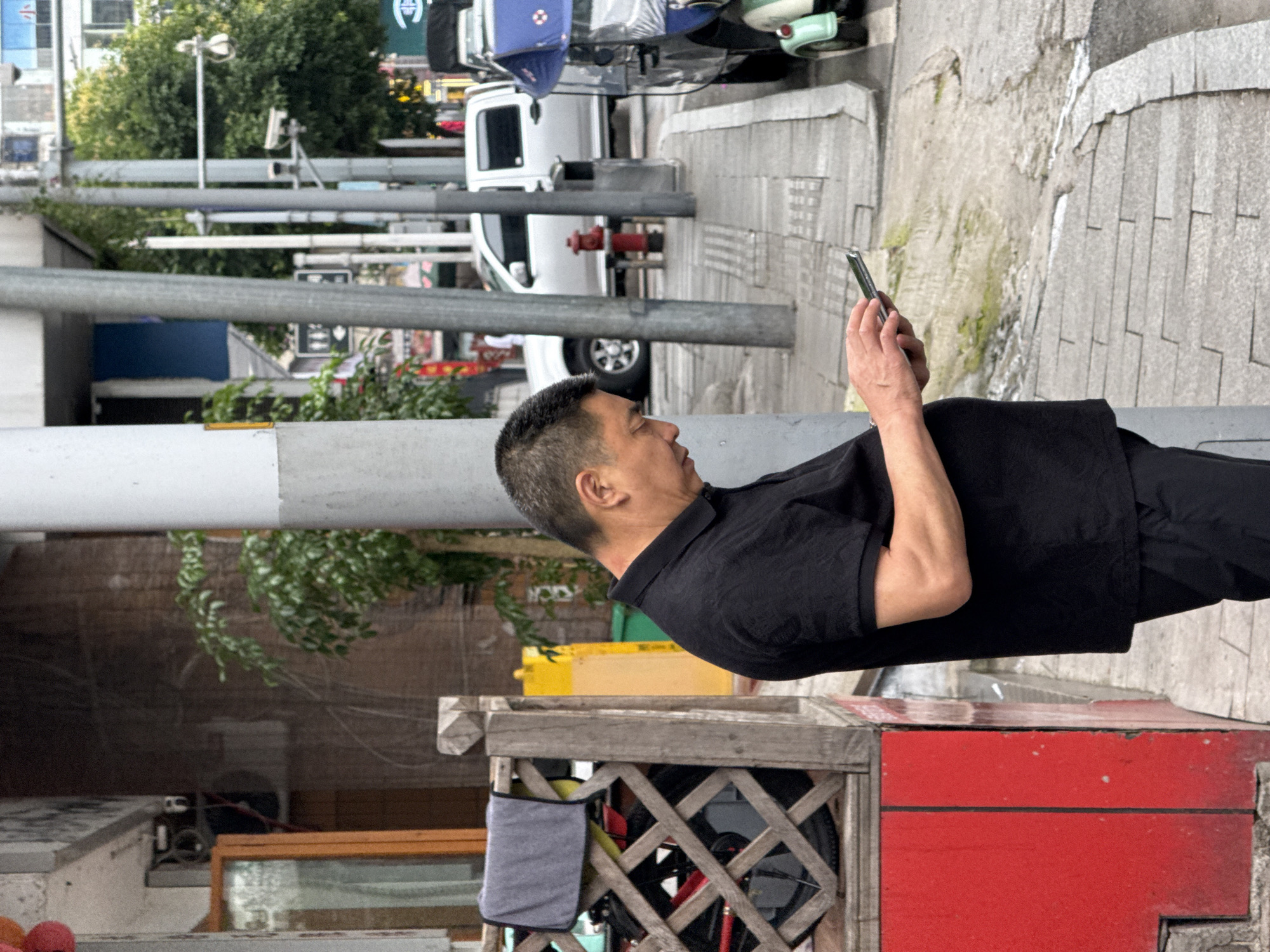 The image shows a man standing on a sidewalk in Jiangjin, China. He is wearing a black shirt and pants and is holding a smartphone, appearing to be taking a photo or video. Behind him, there is a red and wooden structure that appears to be some sort of public furniture or decorative fixture. Further in the background are buildings, traffic lights, street signs, vehicles including vans, and a fire hydrant, indicating an urban environment. The sidewalk is made of concrete and has cracks and wear. The image has been rotated.