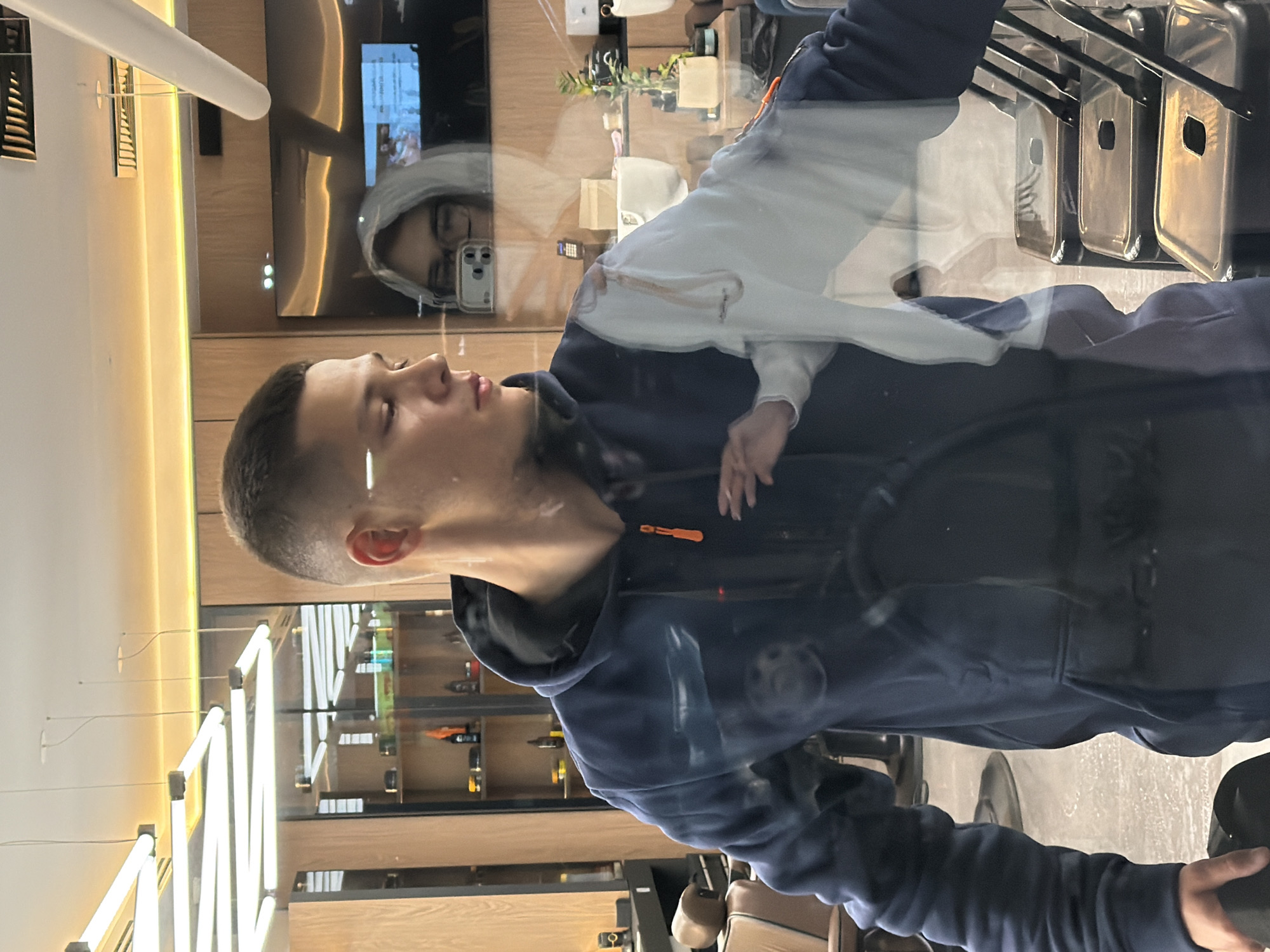 A young man is reflected in a mirror in what appears to be a barber shop. He is wearing a dark blue hoodie with an orange zipper. The mirror reflects a woman in a white hoodie and glasses holding a phone. The barber shop has modern lighting fixtures, wooden paneling, shelves stocked with products, and chairs. The floor is tiled and there is a television screen in the background showing an image. The setting is in the city of Sofia, Bulgaria.