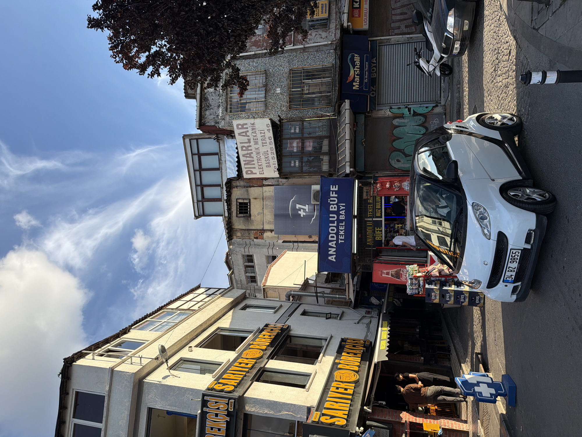 The image depicts a street scene in Istanbul, Turkey, likely in a commercial area. In the foreground, a white Smart car with the license plate "34 BZ 9855" is parked on a cobblestone street. To the left of the car, a blue and white cross-shaped sign stands on the sidewalk. Further back, a row of shops and buildings lines the street. Prominent signage includes "SARIYER BÖREKÇİSİ" displayed on multiple awnings, indicating a bakery or food establishment. Another sign reads "ANADOLU BUFE TEKEL BAYİ," suggesting a buffet and tobacco shop. Above, various signs are visible, including "PINARLAR ELEKTRONIK MEKANIK BASKÜL TERAZİ" which appears to be an electronics and scale repair shop. People are seen walking and standing outside the establishments. In the background, more buildings of varying architectural styles are visible, some appearing older and somewhat dilapidated. The sky above is blue with wispy white clouds. A dark-leaved tree is on the right side of the frame. The overall impression is of a bustling urban environment with a mix of old and new architecture.