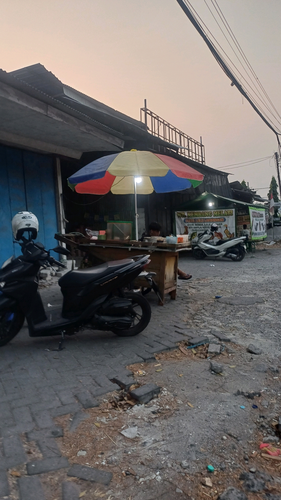 The image depicts a street scene in Klangonan, Indonesia, likely during dusk. A food stall with a colorful umbrella is set up on the side of the road. The stall features a wooden counter with various food items and containers. Two people are visible at the stall, one appearing to be preparing or serving food, and another person seated further back. In the foreground, a black motorcycle is parked, with a white helmet placed on its handlebars. Behind the stall, another white motorcycle is parked. A sign above the stall reads "WR. KEMBANG MELATI PEBEL LELE LAMONGAN" and lists various food items. The ground is paved with bricks in some areas and is uneven and dusty in others, with debris scattered around. The background shows a building with a corrugated metal roof and blue doors. Overhead power lines crisscross the sky, which is a pale orange color, indicating the time of day.