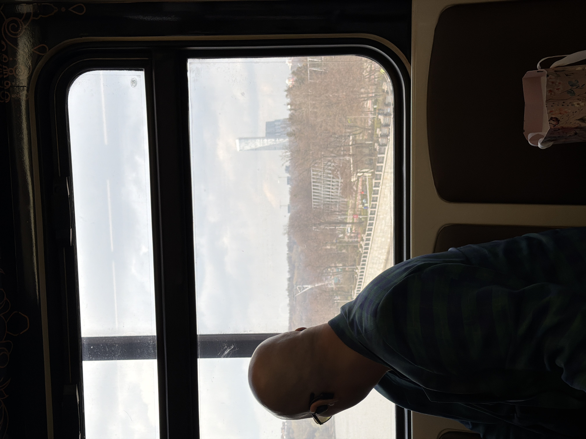 A passenger, possibly a male, is looking out the window of a train. The view from the train window shows a city, likely Moscow, Russia, given the architectural style of the building, landscape, and general atmosphere. The building in the distance is a skyscraper. The background shows trees, possibly in a park. The lighting in the scene is soft, indicating daytime. The train is in motion. The passenger is wearing glasses.