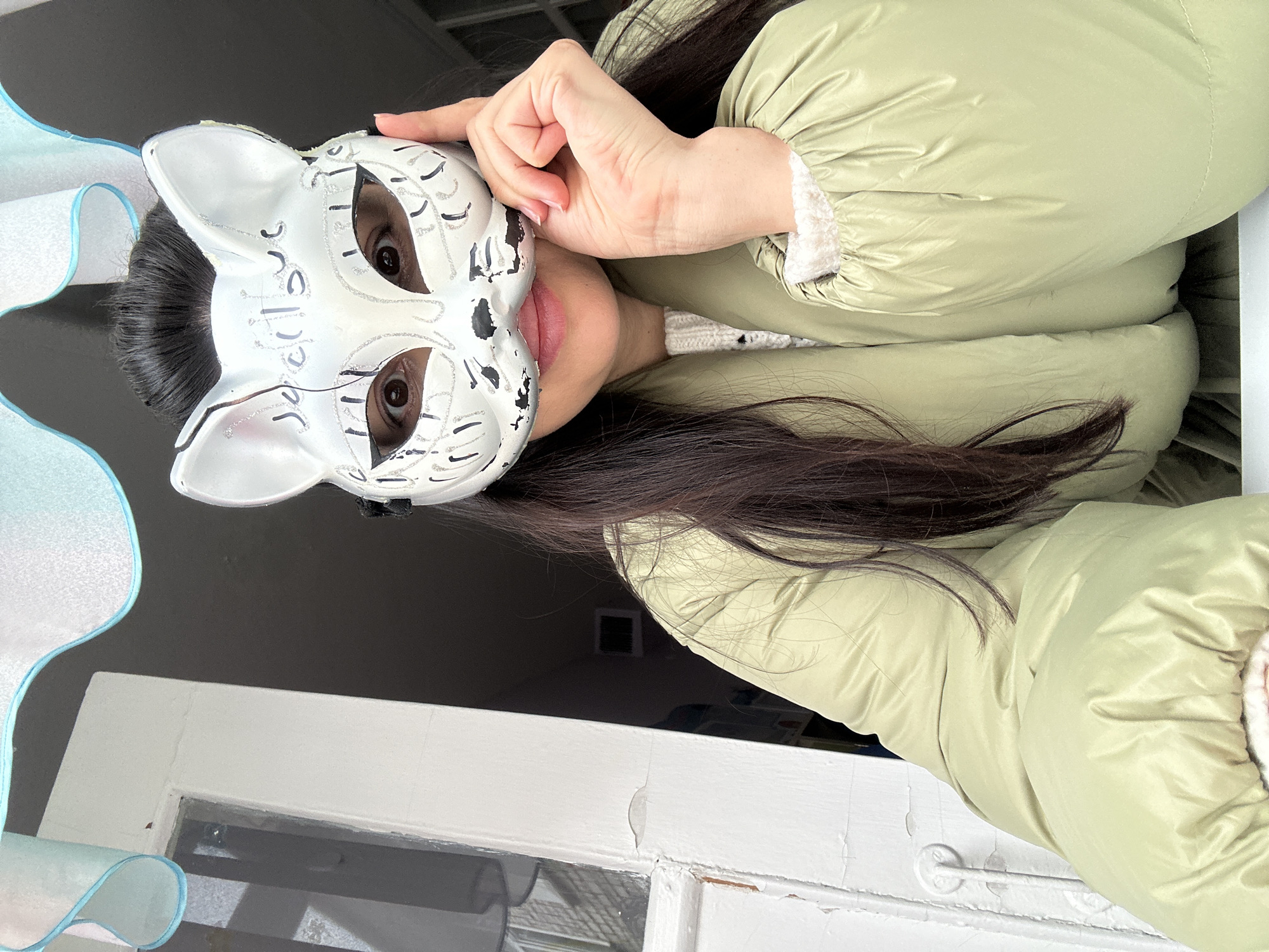 A person wearing a light green puffer jacket and a white cat mask is visible. The person is standing near a white window frame with peeling paint. The location is Chaikovsky, Russia.