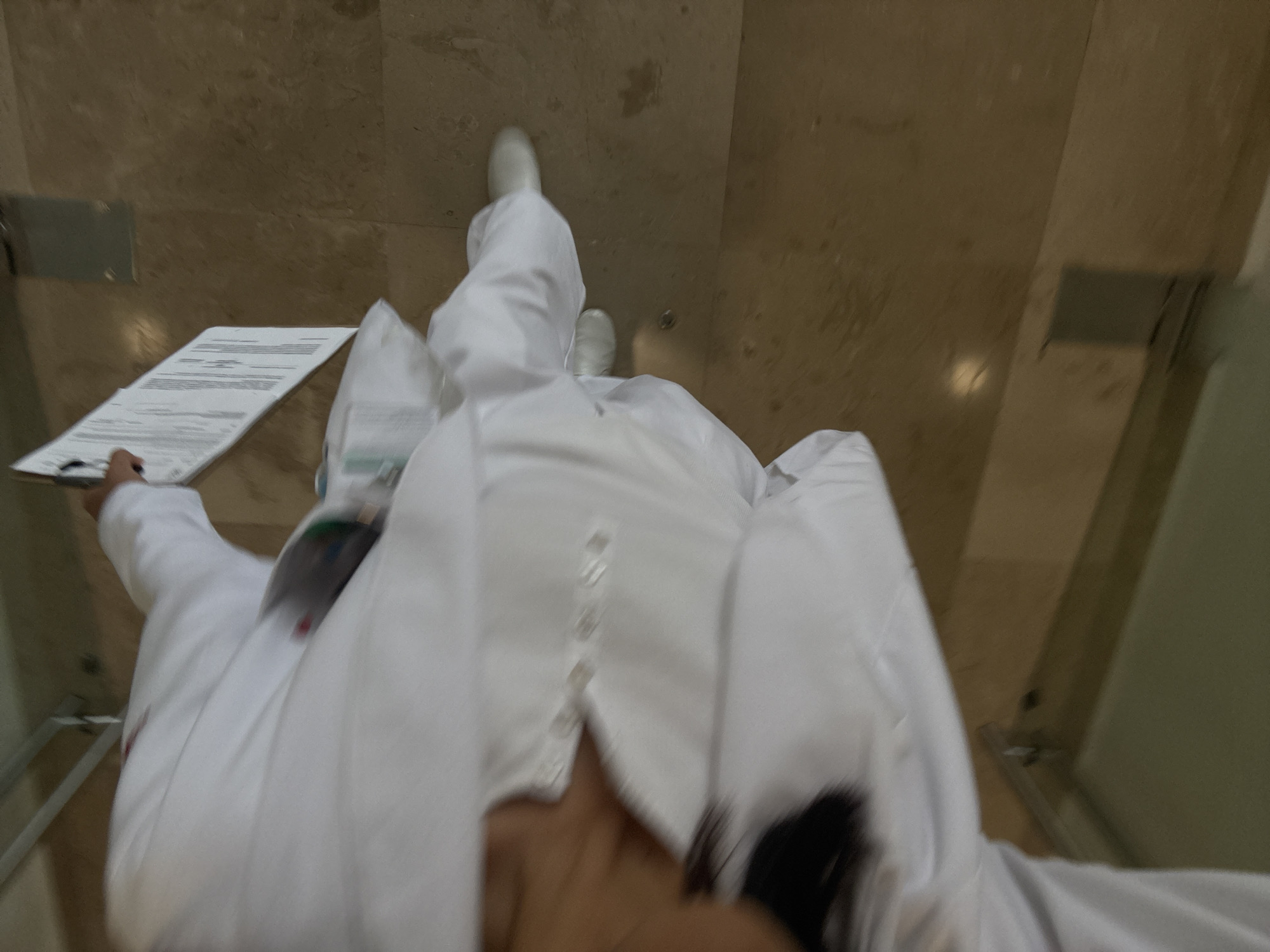 A first-person perspective photograph shows a person wearing white medical scrubs and white shoes walking down a corridor. The person is holding a clipboard with papers in their left hand. The floor is tiled in a light brown color. It is not possible to determine the location in Puebla, Mexico from the visual information.