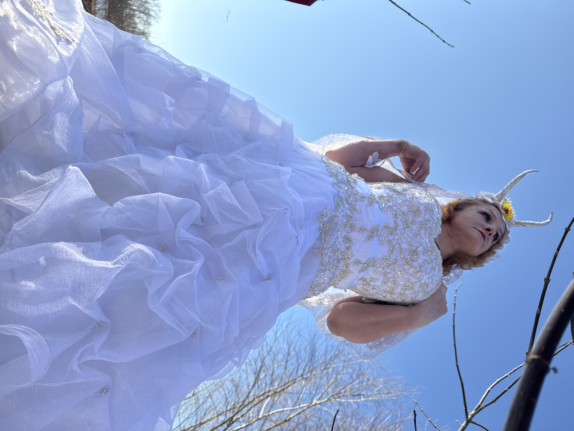 A woman in a white dress with gold embellishments poses in an outdoor setting under a bright blue sky. She wears a headdress with white horns and a yellow flower. The dress has a layered and ruffled skirt. Tree branches without leaves are visible in the foreground and background. The location appears to be a natural area, possibly a park or forest. While the image is possibly taken in Chaikovsky, Russia, there is not enough information to confirm the location.