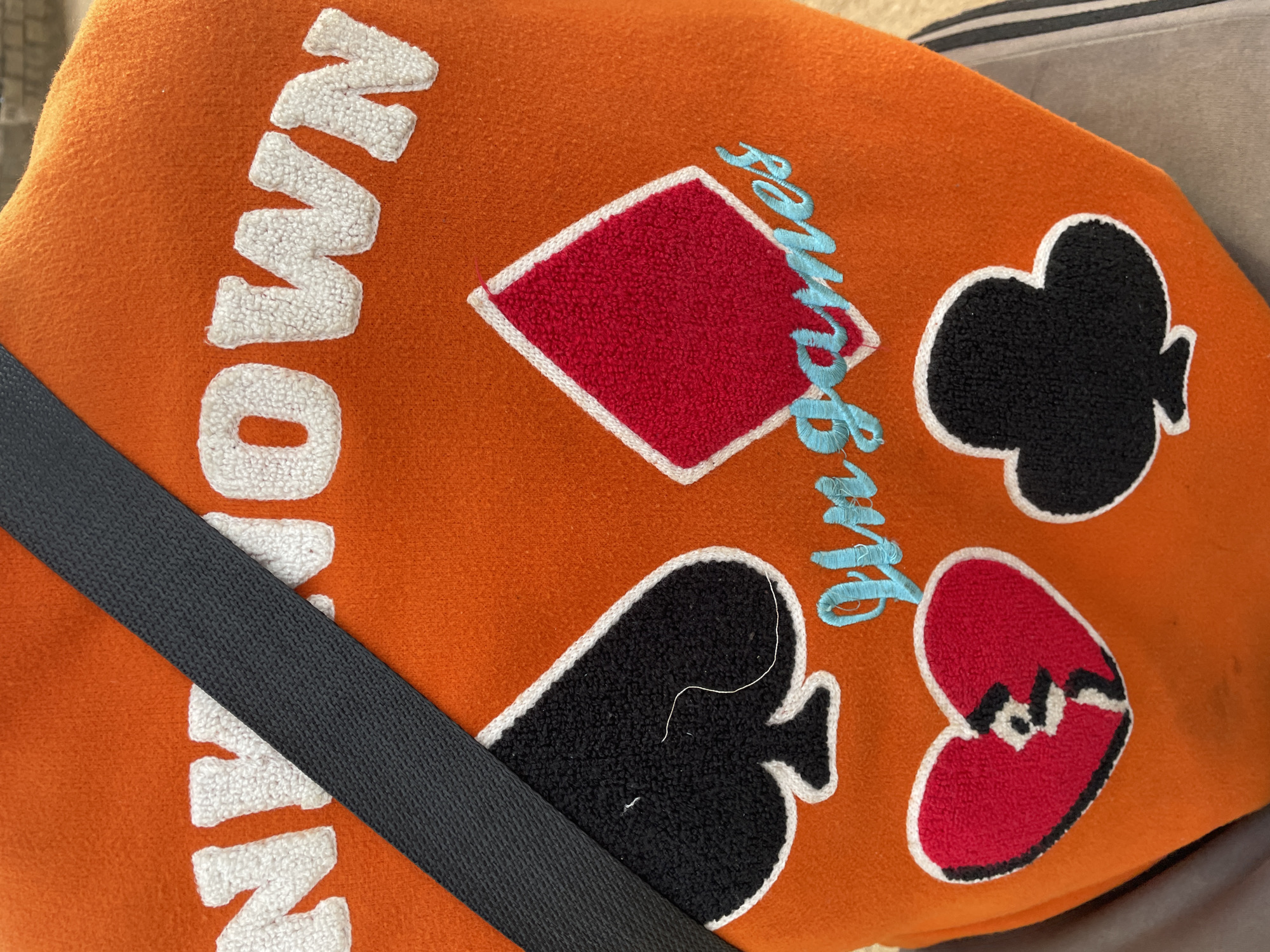 The image shows a close-up of an orange, textured fabric bag with a black strap crossing diagonally. The bag is decorated with playing card suit symbols – a red diamond, a black club, a black spade, and a red heart with a black crack – each outlined in white. "Monopoly Games" is embroidered on the bag in white and blue thread. The location in the city of Gura Top, Nigeria is unknown.