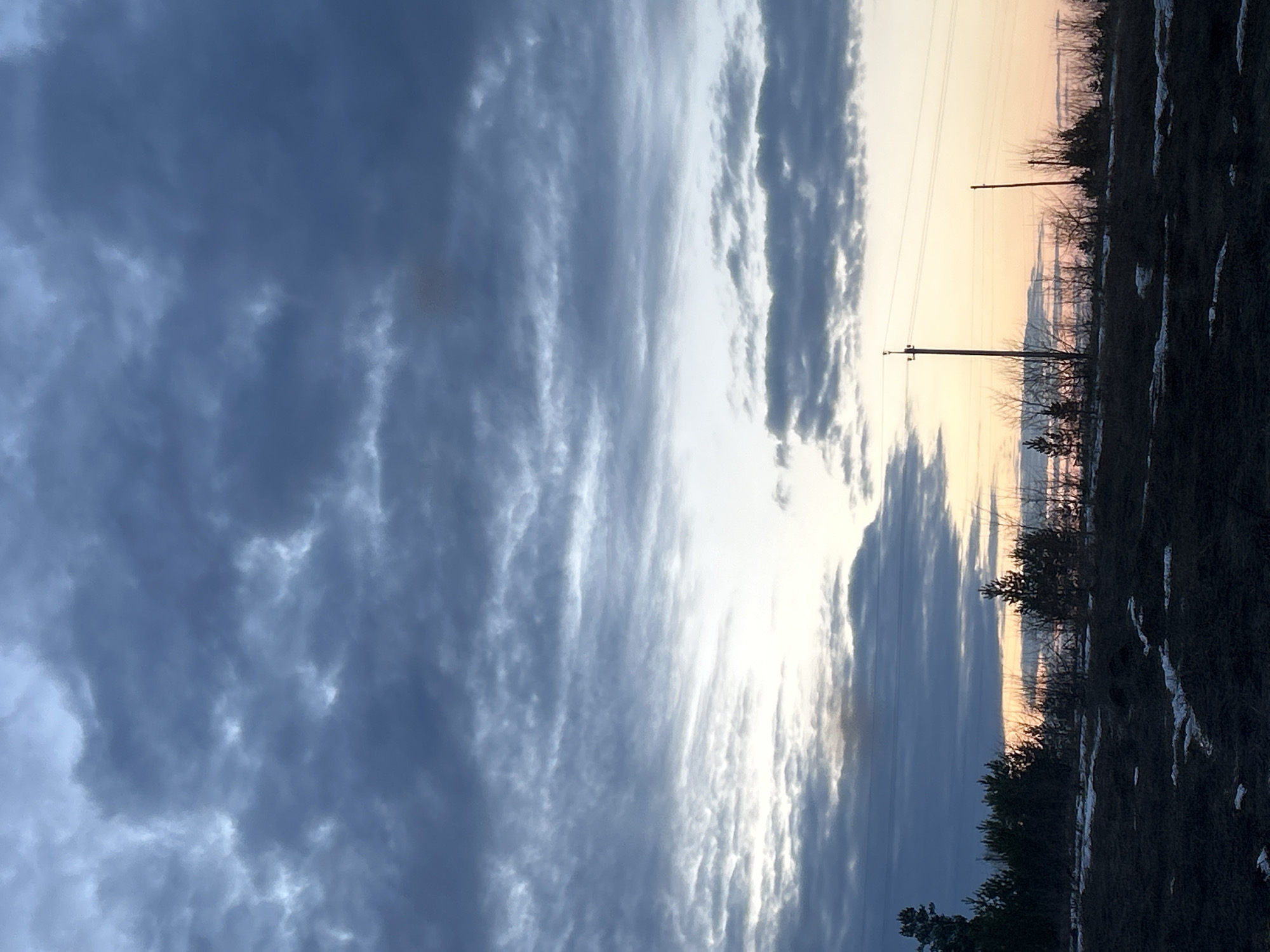 The image shows a landscape scene at dusk or dawn. The sky is dominated by clouds with varying shades of blue and white, suggesting a dramatic weather condition. The horizon reveals a pale orange glow, possibly indicating sunrise or sunset. Two utility poles with power lines are visible against the sky. The foreground shows an area of land with dark vegetation and patches of snow. The precise location in Фоки, Russia cannot be determined from this image alone.