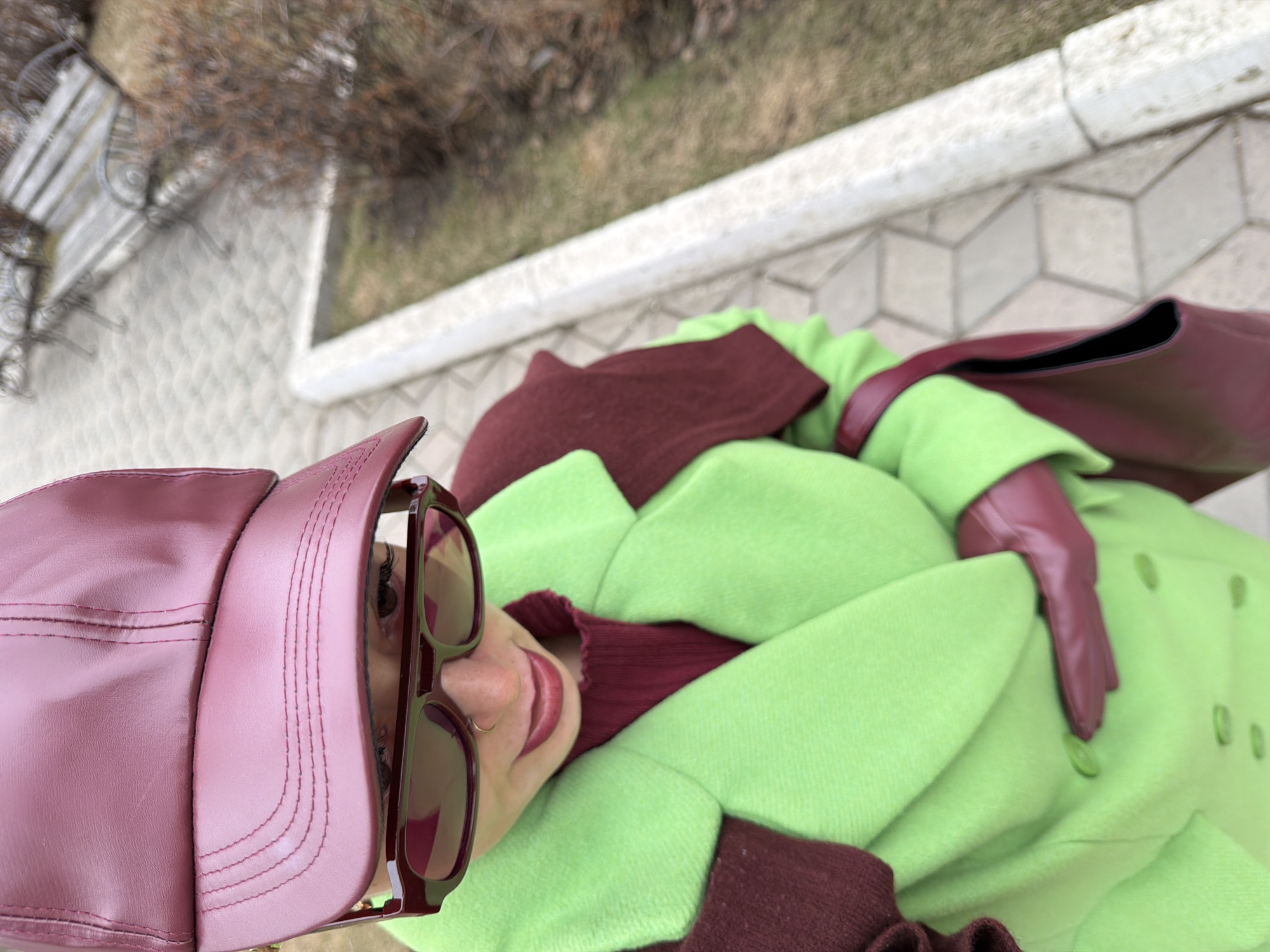 A medium-shot, high-angle, color photograph depicts a person wearing a burgundy leather cap, matching sunglasses, a green wool coat with burgundy accents, and burgundy leather gloves. The person is laying on a paved surface consisting of a repeating pattern of geometric shapes. A bench can be seen in the upper-left corner. In the background, blurred trees suggest an outdoor, urban environment, possibly within the city of Krasnoyarsk, Russia.