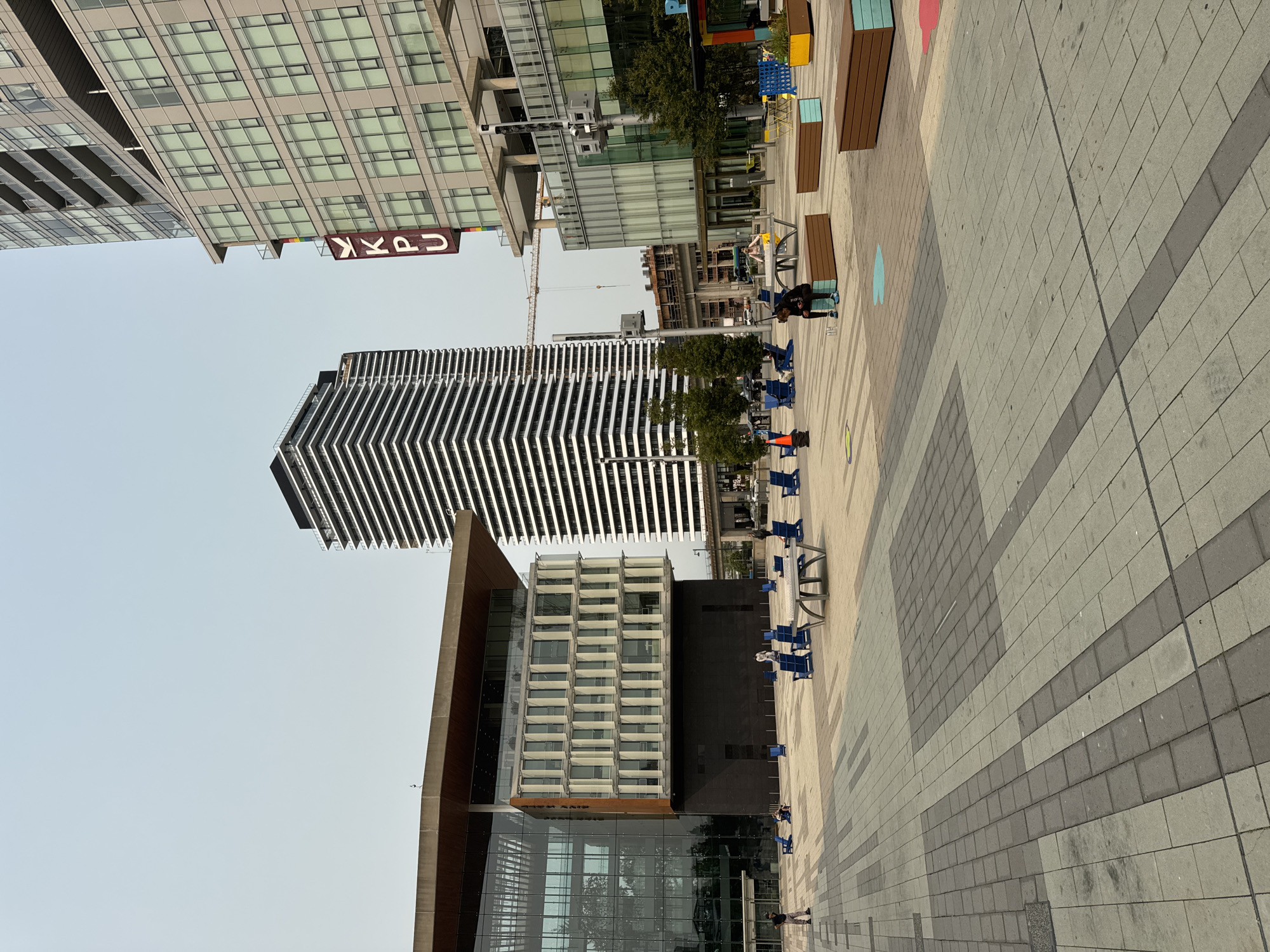 The image captures a wide outdoor plaza in Surrey, Canada, likely part of a contemporary urban development. In the foreground, a patterned pavement made of large rectangular grey and beige tiles stretches across the scene. Several blue Adirondack chairs are scattered across the plaza, some occupied by individuals. A metal picnic table with bench seating sits centrally. Further back, long, low brown wooden benches with light blue accents are placed on the pavement, with some individuals seated on them. Colorful planters are visible, one with a yellow and blue design and another with a red splash of color.
Dominating the background are modern buildings. To the left, a large glass-fronted building with a concrete facade and an overhanging brown roof is prominent. To the right and center, a tall, sleek skyscraper with a black and white striped facade is under construction, with a crane visible above it. Adjacent to this skyscraper is another multi-story building with a light grey facade and numerous windows. A vertical sign with the letters "KPU" in red lettering is attached to this building. The sky is hazy and overcast. The scene appears to be a public space designed for leisure and gathering, integrated within a growing urban environment.