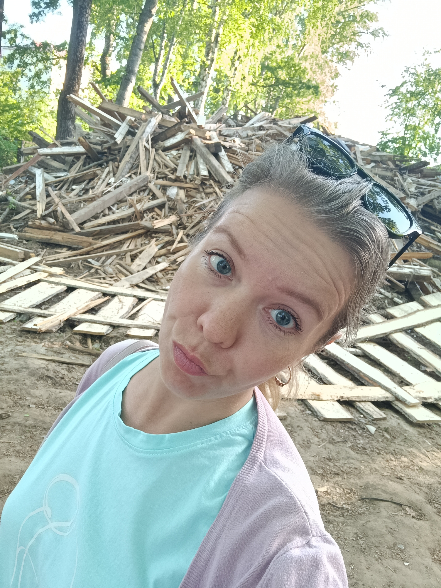 A selfie of a woman with fair skin, blue eyes, and freckles. She wears a light blue t-shirt and a light purple cardigan. Sunglasses rest on top of her head. She is positioned in front of a large pile of wooden debris, potentially demolition materials. The background includes trees suggesting an outdoor location. Given the prompt, the location is likely in the city of Chaikovsky, Russia.