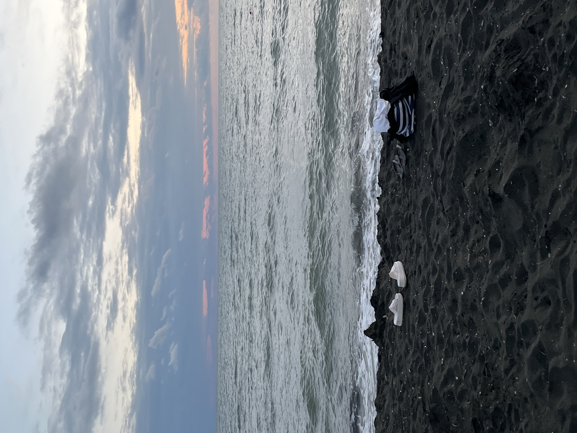 This media file depicts a beach scene at Ureki, Georgia. The image is oriented vertically, with the sky occupying the left portion and the sea and beach on the right.
The sky is filled with dramatic clouds, with streaks of orange and pink indicating either sunrise or sunset. The sea is a muted gray-green, with gentle waves lapping at the shore. The beach is composed of dark, fine sand, dotted with small shells.
On the sand, near the water's edge, are several objects. A striped garment, possibly a shirt or towel, is folded alongside a white item, perhaps another piece of clothing. Further up the beach, a pair of white shoes, possibly sneakers or slip-ons, are placed side by side. The scene suggests a moment of pause, perhaps someone has recently been swimming or is preparing to leave the beach.
The time of day is likely early morning or late evening, given the colors in the sky. The weather appears calm. The dark sand is a distinctive feature of Ureki, known for its magnetic sand. The overall mood is serene and quiet, with a touch of natural beauty highlighted by the dramatic sky and the minimalist arrangement of objects on the beach. No visible text is present in the image.