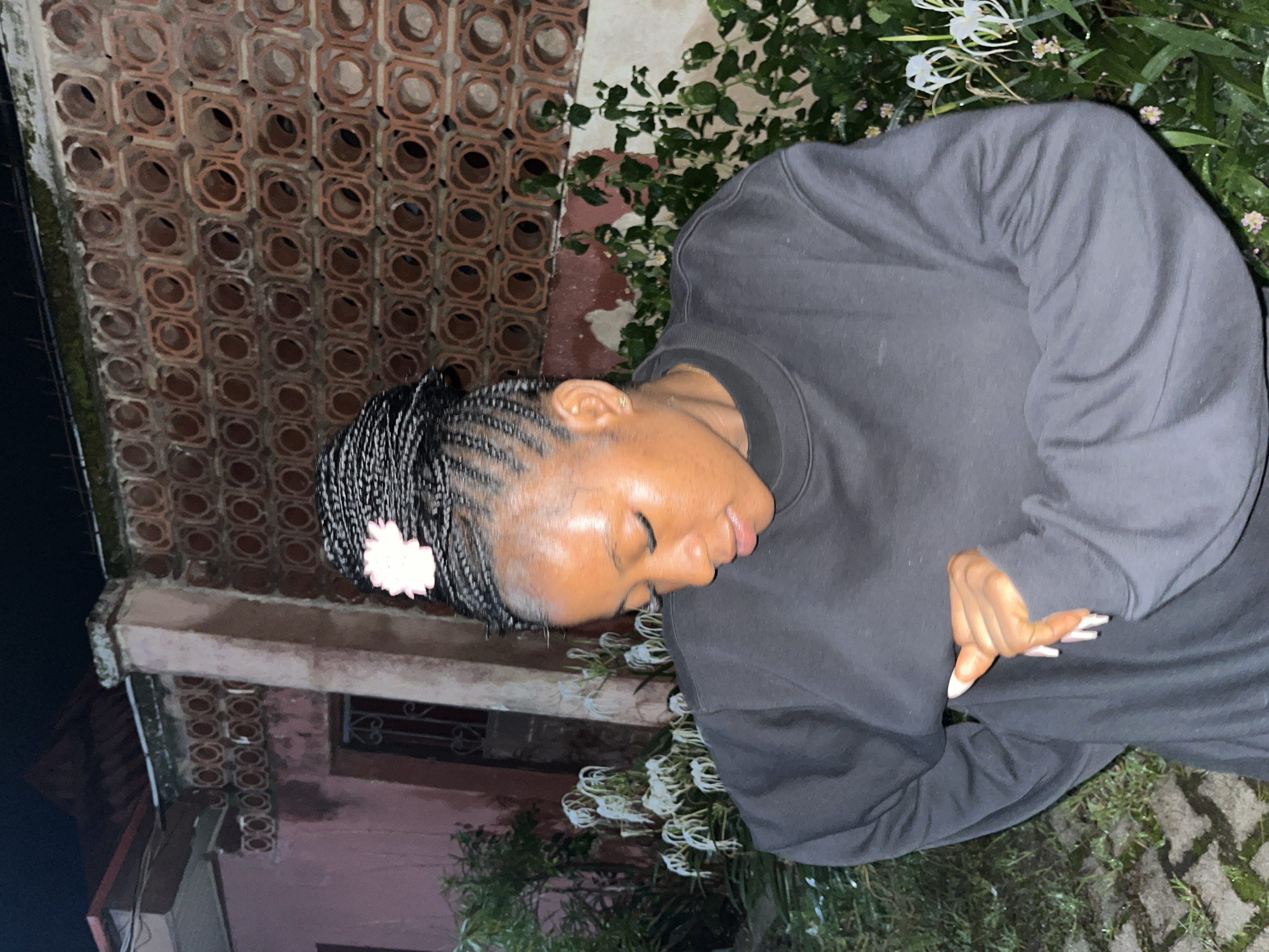 In an outdoor setting at night, a young woman with braided hair adorned with a pink flower is captured from the chest up, looking down and to her right. She is wearing a dark, long-sleeved sweatshirt. Her left hand is visible, resting near her chest, with manicured fingernails. The background features a textured wall constructed from patterned terracotta bricks, a pink-hued building with a red-tiled roof, and lush green foliage with white flowers. The ground appears to be paved with interlocking stones. The scene suggests a residential area, potentially within the city of Jos, Nigeria, given the architectural style and vegetation.