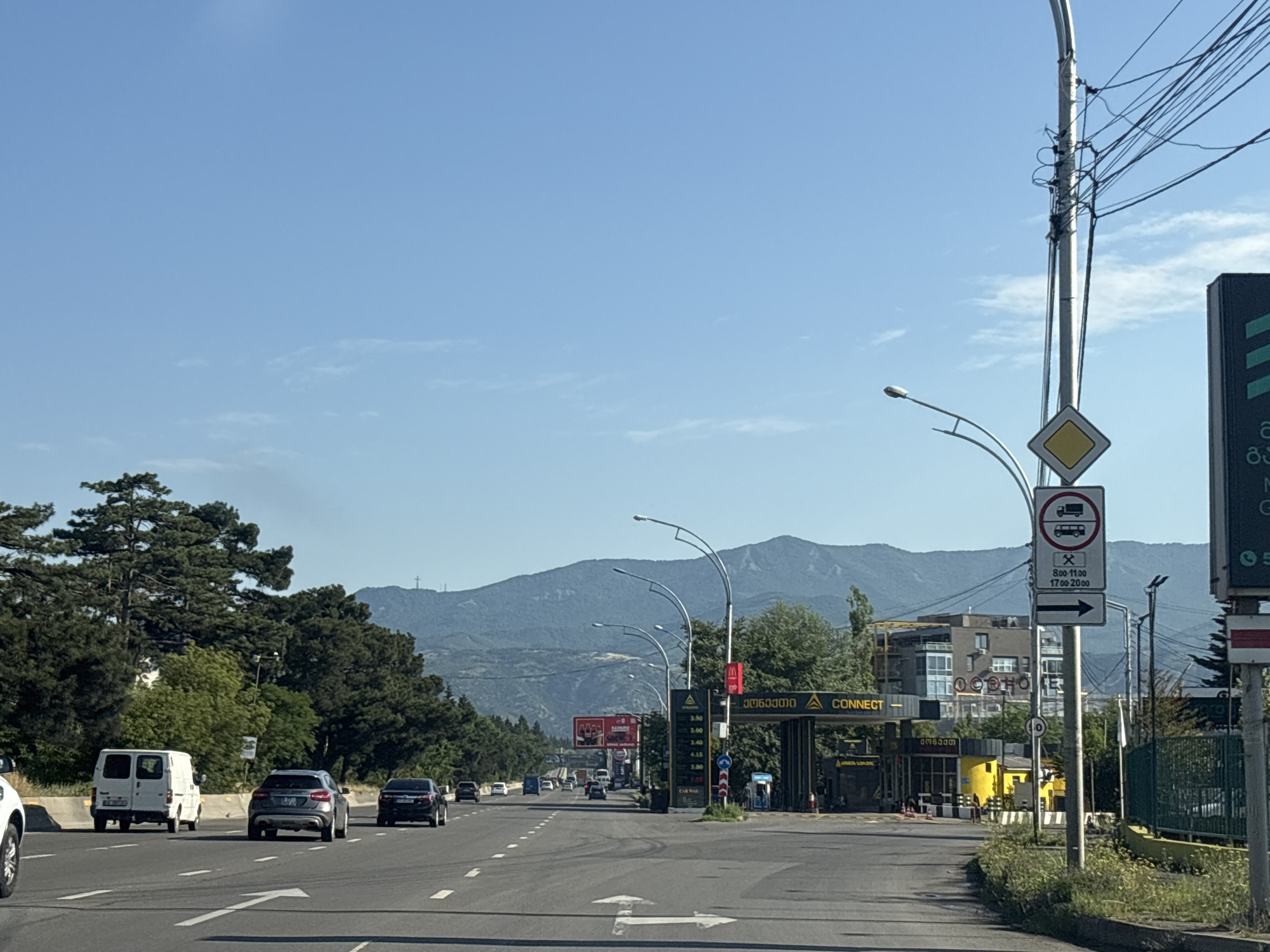 The image depicts a street scene in Tbilisi, Georgia, on a clear day with a blue sky. In the foreground, a paved road with white lane markings is visible, with several cars driving on it. To the left, a white van is driving in the left lane, followed by a gray SUV and a black sedan in the right lane. Further down the road, more cars are visible.
On the right side of the road, a traffic light pole with numerous wires is prominent. A diamond-shaped yellow sign with a red border and a black truck symbol is mounted on the pole, indicating a restriction for trucks. Below it, a sign with a pickaxe and hammer symbol and times "8:00-11:00" and "17:00-20:00" suggests specific hours for a task or service. A white directional arrow pointing to the right is also present on the pole.
Beyond the road, there is a yellow structure with the word "CONNECT" written in large letters, appearing to be a toll booth or service area. Signs with Georgian text are also visible on the structure. Green trees line the sides of the road and the background. In the distance, a range of mountains under a hazy sky can be seen.
A large billboard with red and white lettering is situated behind the trees on the left side of the road. A McDonald's sign is visible on a yellow and black structure on the right. A tall, dark green fence runs along the right edge of the road in the mid-ground. The overall scene suggests an urban or suburban environment with ongoing traffic.