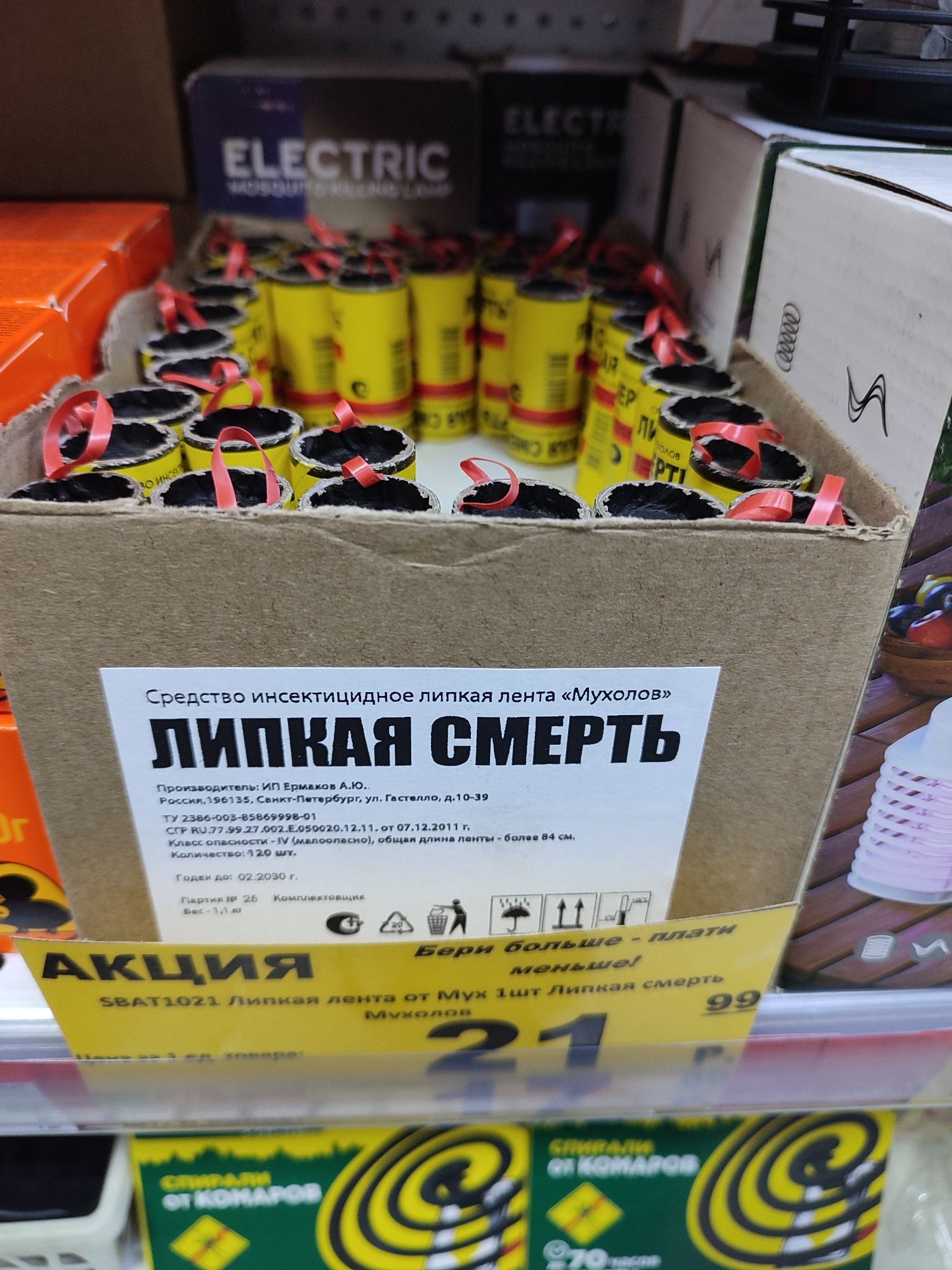A close-up, eye-level, indoor shot captures a display of insect repellent products in a retail environment. In the foreground, a cardboard box contains numerous yellow cylinders with red ribbons, presumably insect repellent devices. A white label on the box reads "Средство инсектицидное липкая лента «Мухолов»" (Insecticidal sticky tape "Flycatcher") and "ЛИПКАЯ СМЕРТЬ" (Sticky Death). Below this, manufacturing details and product information are provided, including "Производитель: ИП Ермаков А.Ю." (Manufacturer: IP Ermakov A.Yu.) and "Россия, 196135, Санкт-Петербург, ул. Гастелло, д.10-39" (Russia, 196135, Saint Petersburg, Gastello St., 10-39). Further down the label, the quantity is listed as "Количество: 120 шт." (Quantity: 120 pcs.). A yellow angled promotional banner at the bottom of the box declares "АКЦИЯ" (Promotion) and mentions "SBAT1021 Липкая лента от Мух 1шт Липкая смерть" (SBAT1021 Sticky tape for flies 1pc Sticky Death) with prices of "21" and "99".
To the right of the cardboard box, a stack of pink plastic items, possibly more insect repellent devices or containers, is partially visible. In the background, shelves are stacked with boxes of various sizes and colors, including some with the word "ELECTRIC" printed on them, suggesting other electronic or pest control products. The lighting appears to be artificial, typical of a store. The scene is static, with no people or actions explicitly depicted. The overall impression is of a commercial display promoting pest control solutions.
