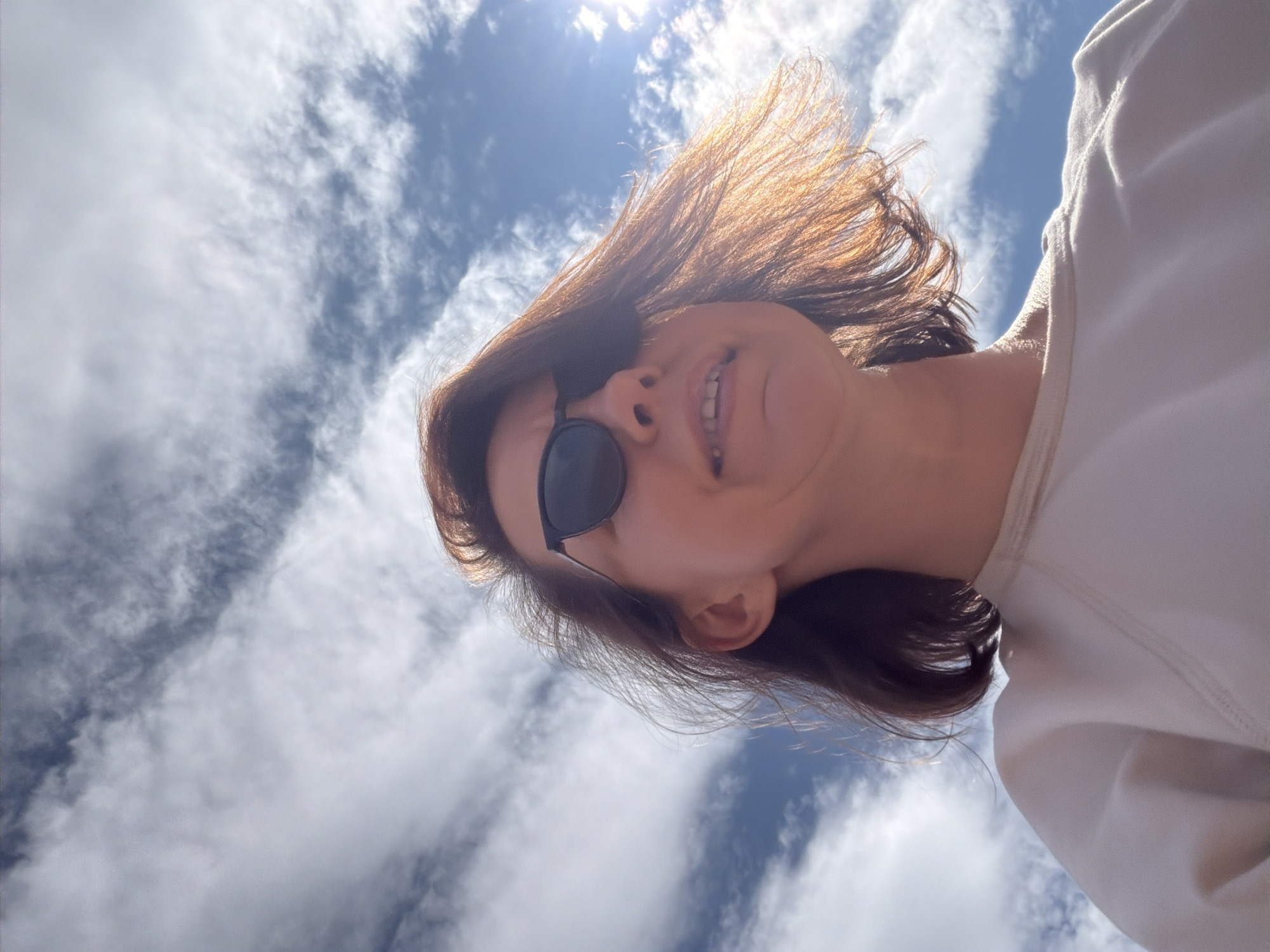 A woman wearing sunglasses and a light-colored sweatshirt looks up towards a bright, cloudy sky. The sun is visible, casting a warm glow on her hair. The image appears to be taken outdoors, possibly in Chaikovsky, Russia, given the context.