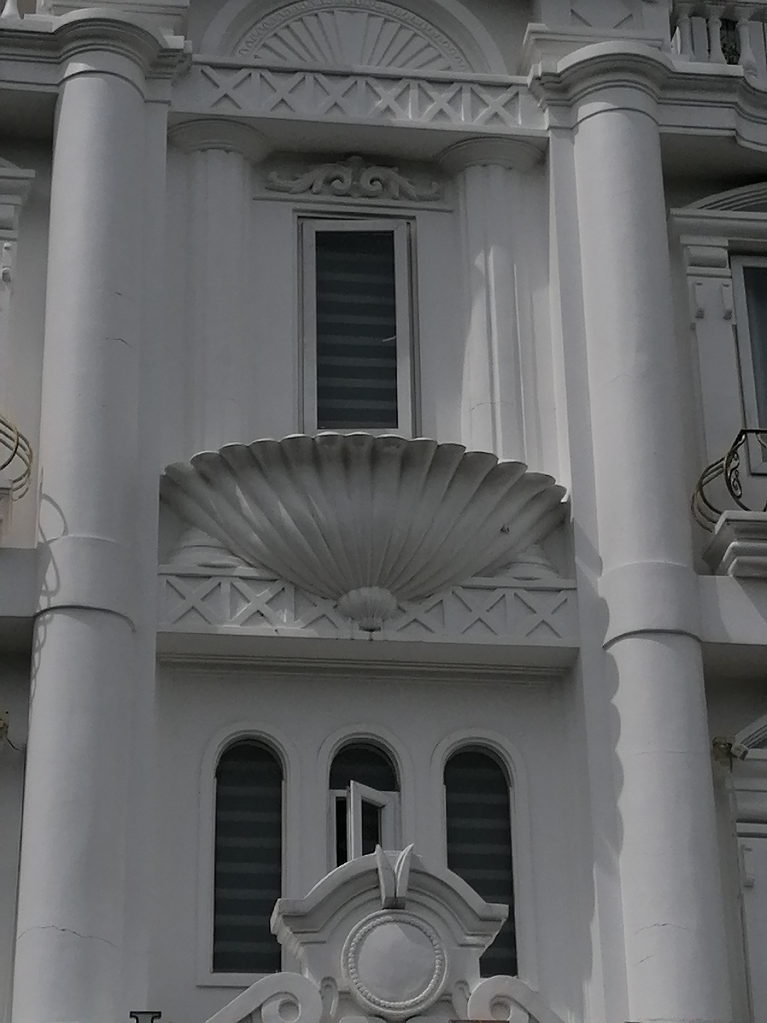 The image shows a close-up, eye-level view of the facade of a white, neoclassical-style building in Da Lat, Vietnam. The building features prominent fluted columns, arched windows with horizontal blinds, and decorative architectural elements such as a large seashell-shaped motif above a lower row of windows. The building appears to be in good condition, though some minor cracks are visible on the exterior paint. The lighting suggests it is daytime, with shadows indicating the sun is at an angle. No people or specific activities are discernible in the image.