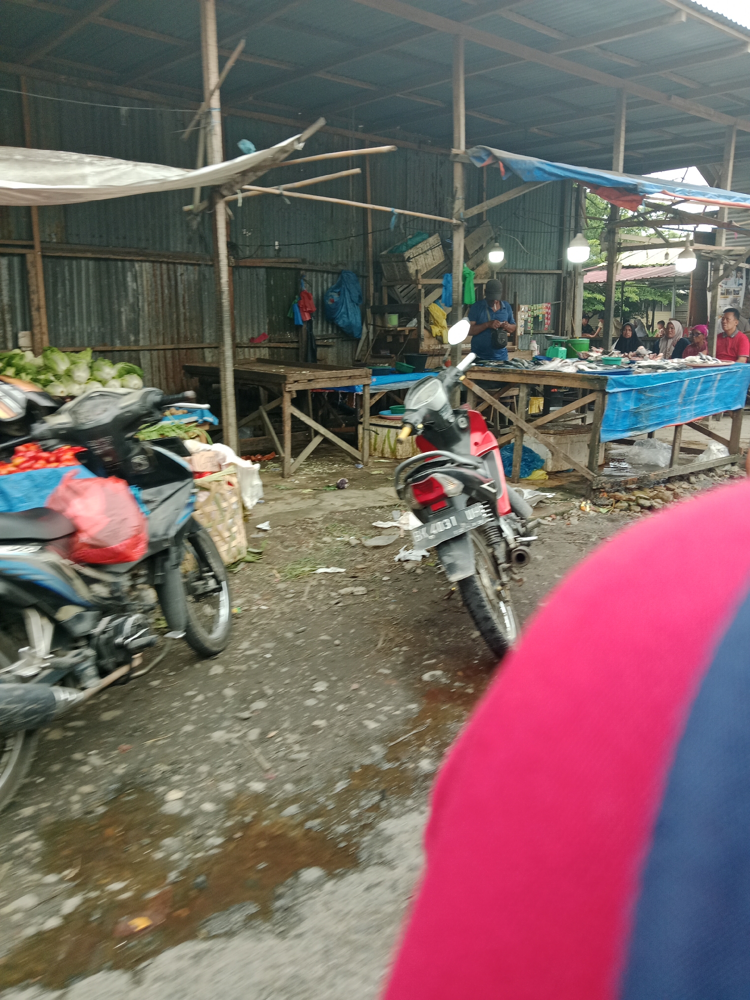 This image depicts an outdoor market scene in the City of Binjai, Indonesia. The market appears to be partially covered by a corrugated metal roof supported by wooden beams. Tarpaulins of various colors, including white, blue, and red, are used as additional roofing and coverings.
In the foreground, two motorcycles are parked. The motorcycle on the left is partially visible, showing a blue and black color scheme with a red bag tied to its rear. A pile of red tomatoes is also visible in front of this motorcycle. The motorcycle on the right is a red and black model with a license plate that reads "BK 4031 UP".
The market stalls are constructed with wooden planks. One stall in the middle ground displays a variety of fish laid out on a blue tarpaulin. Several people are gathered around this stall, appearing to be customers or vendors. The people include women wearing headscarves and men.
Behind the stalls, there are stacks of crates and other market goods. Green vegetables, possibly cabbage or lettuce, are piled up on the left side of the image. The ground is uneven and appears to be a mixture of dirt and gravel, with some puddles of water.
In the background, the market extends further with more stalls and structures. The overall impression is a bustling, open-air market environment.