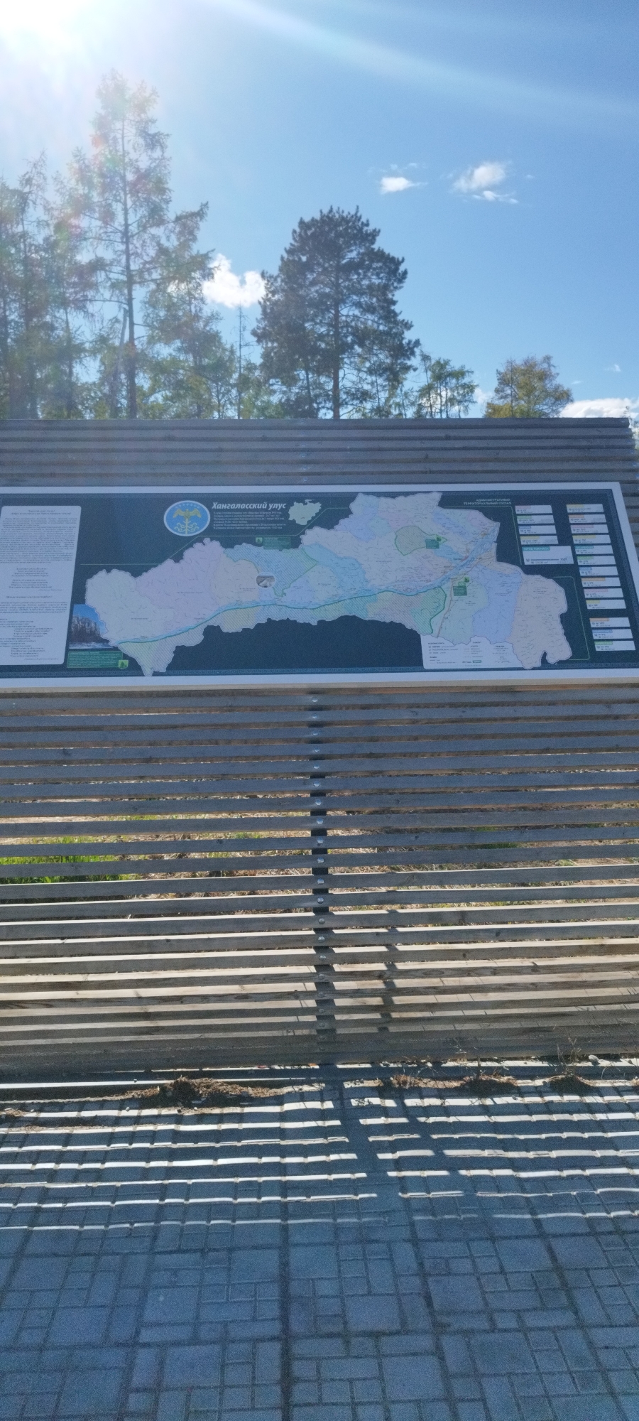 The image shows a sign with a map of "Kangalassky ulus" (Хангаласский улус) in Tekhtiur, Russia, viewed through a wooden slatted fence. The sky is blue with some clouds and sunlight. Trees are visible in the background. The ground is paved with rectangular bricks, casting shadows from the fence. No people are visible.