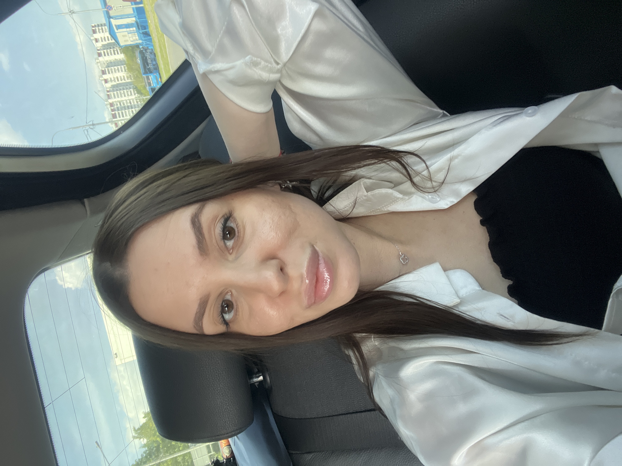A selfie of a woman with light brown hair wearing a white shirt and a black top in the backseat of a car. The car's window shows a building and trees in the background, suggesting an urban environment. The image does not provide sufficient information to determine if it was taken in Moscow, Russia.