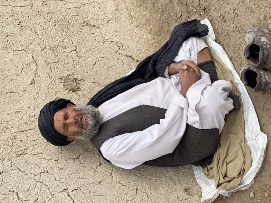 An elderly man with a gray beard and a black turban lies on the ground, dressed in a white shirt, dark vest, and light-colored pants. He is positioned on a white and beige cloth, with his arms wrapped around his legs. A dark cloth drapes over part of his body. Nearby, a pair of worn shoes sit on the ground. The ground's texture is rough and cracked.