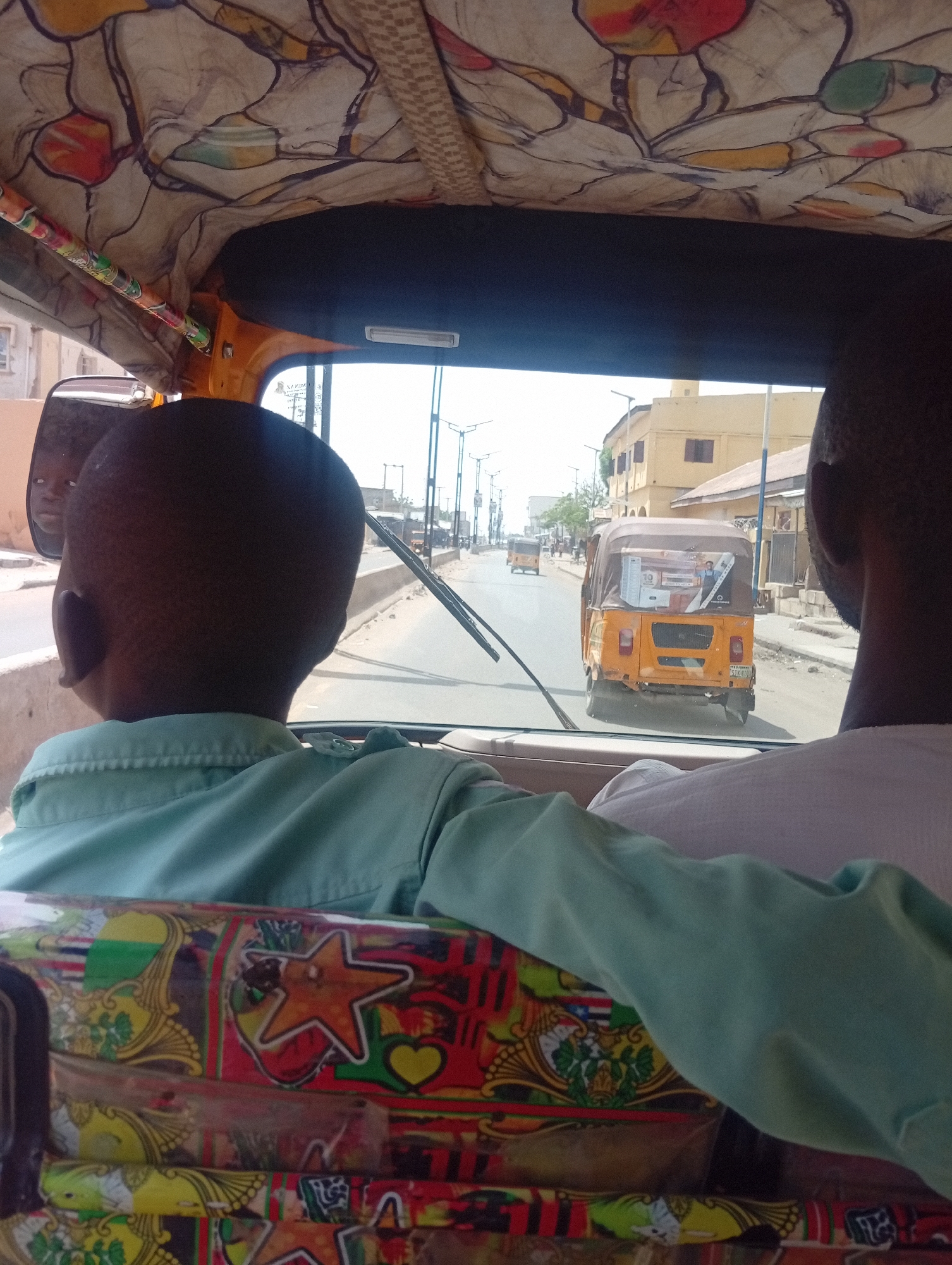 View from inside a motorized tricycle taxi, known as a "keke," in Gama, Nigeria. Two male passengers are visible in the foreground; one is a child with a light green shirt on the left, and the other, on the right, has a white shirt. Both are facing forward. Another keke is in front of them on the road. Buildings and streetlights are visible along the road. The interior of the keke features patterned fabric on the roof and decorated bars. The keke in front has advertisements on its back.