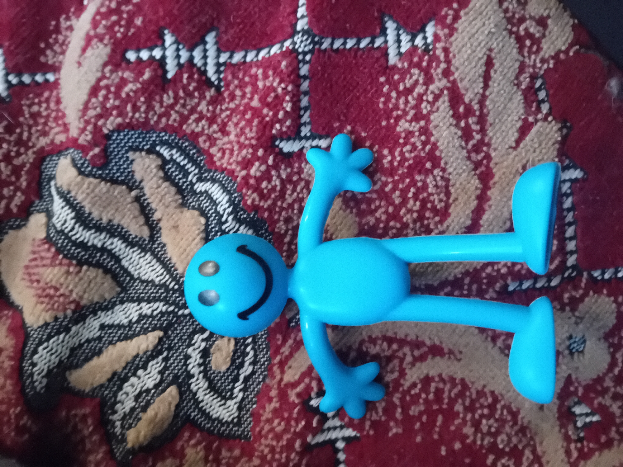 A close-up, overhead shot shows a bright blue, stylized toy figure with a large head, two circular eyes, and a wide smile. The figure has a simple, rounded body, four-fingered hands, and flat, rounded feet. It is standing upright on a textured surface that appears to be a rug with a red background and a patterned design featuring light brown and black elements. The lighting is direct and casts a slight shadow around the figure. The background pattern consists of repeating abstract shapes and lines. The image was captured indoors, suggesting a domestic environment. The specific location in Chuhuiv, Ukraine cannot be determined from the visual information provided.