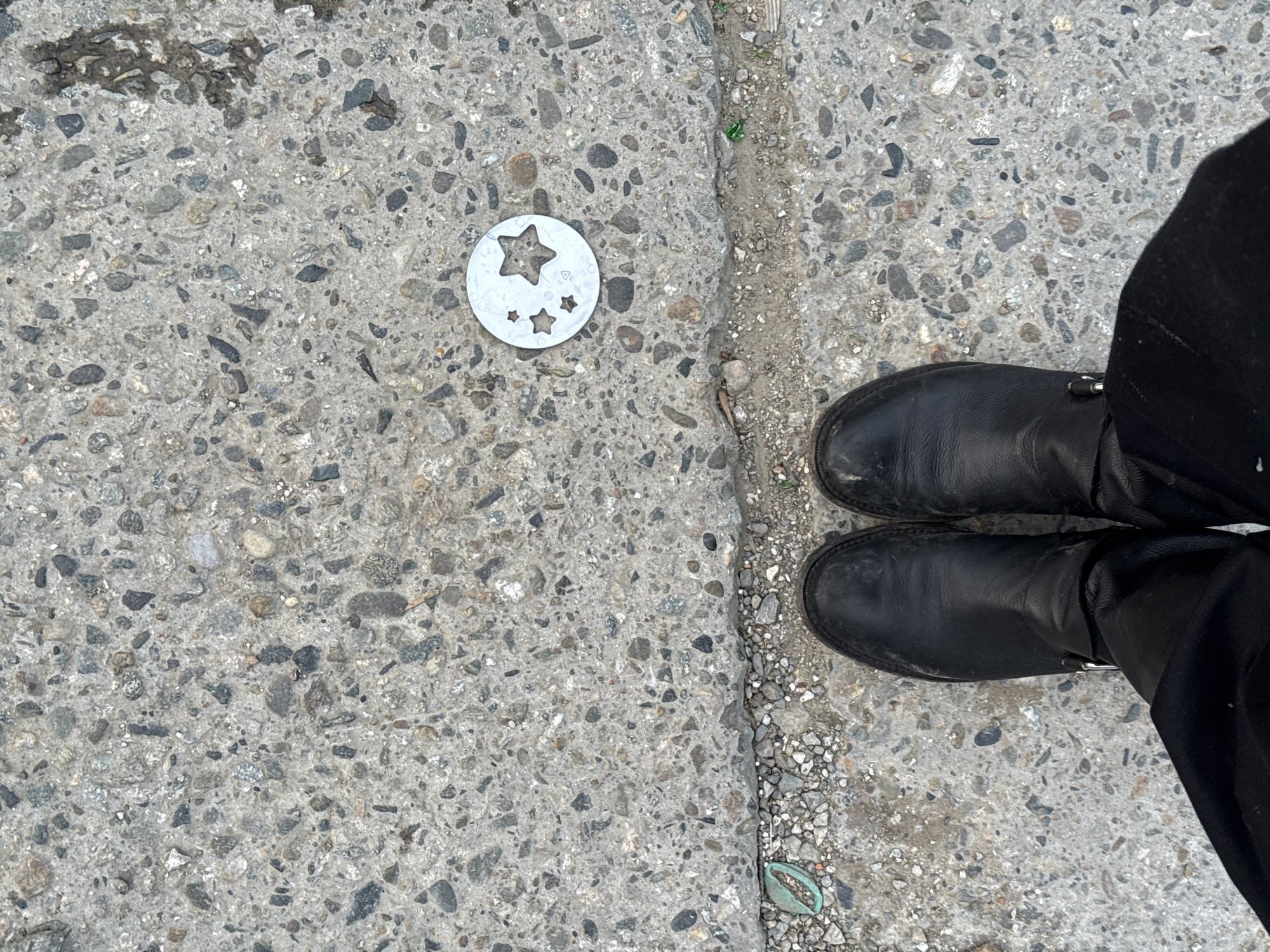 The image shows a section of sidewalk or pavement with an adjacent curb. On the left side of the image, there is a circular metal disc with star-shaped cutouts resting on the pavement. To the right, a person is standing with their feet visible. They are wearing black boots and black pants. The pavement appears to be made of concrete with embedded stones. The edge of the curb is visible, with smaller pebbles and some minimal plant growth along the edge. The location (Sochi, Russia) is not discernible from the image itself.