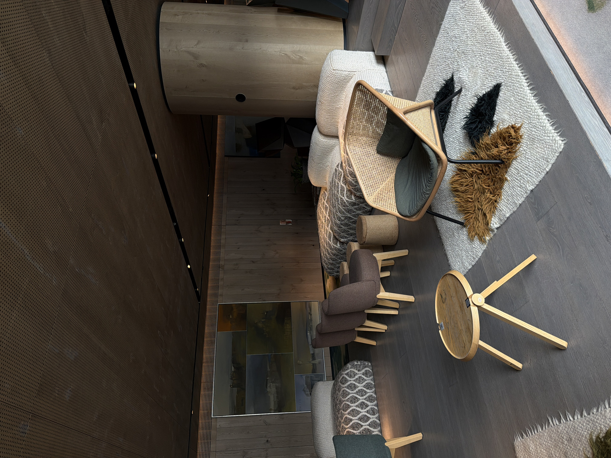 An overhead shot captures a modern, upscale lounge or waiting area, characterized by a sophisticated and warm aesthetic. The dark wood plank flooring runs throughout the space, providing a rich foundation. A prominent dark brown perforated wall, accented with sleek black strips and small embedded lights, stretches across the left side of the frame, curving slightly upwards. In the background, a lighter wood panel wall is visible, complementing the dark flooring.
The lounge is furnished with a variety of contemporary pieces. A large, light-colored textured sofa or bench, possibly modular, is adorned with a woven throw blanket. Adjacent to it, a stylish armchair crafted from light wood and cane webbing, featuring a dark grey cushion, stands out. Underneath this chair lies a shaggy, off-white rug with fringed edges, on which two decorative, shaggy elements—one black and one brown—resemble abstract shapes or animal features.
Further into the room, two dark brown upholstered armchairs with light wooden legs are arranged, positioned near a large, multi-panel framed artwork on the wall. This artwork displays a series of serene landscape or seascape paintings in muted tones. A small, round wooden side table with three splayed legs is placed on the dark flooring, with a tiny, illegible tag visible on its surface. Another small, textured, round stool is also present. The overall lighting appears bright, suggesting a daytime setting or a well-lit interior, creating a tranquil and inviting atmosphere. No people are visible.