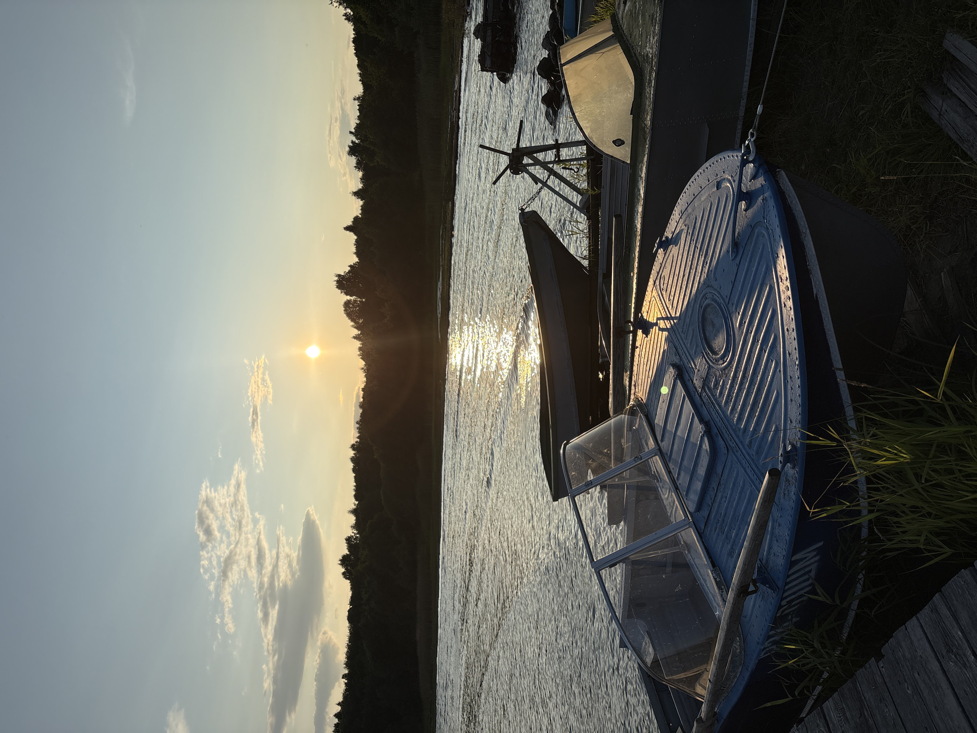 The image depicts a serene sunset over a body of water, likely a river or lake, in Tipinitsy, Russia. Several boats are docked on the shore, with the foreground dominated by a blue motorboat with a clear windshield. Behind it, a larger, darker boat is partially visible, and further back, another boat is propped up on a stand. The sun, a bright orb, is setting low in the sky, casting a warm, golden glow on the water and illuminating the clouds. A dense line of trees forms the horizon, silhouetted against the vibrant sky. The water is rippled, reflecting the light of the setting sun. The scene is peaceful, with no people visible. The environment suggests a rural or natural setting, possibly a marina or a quiet waterfront area.