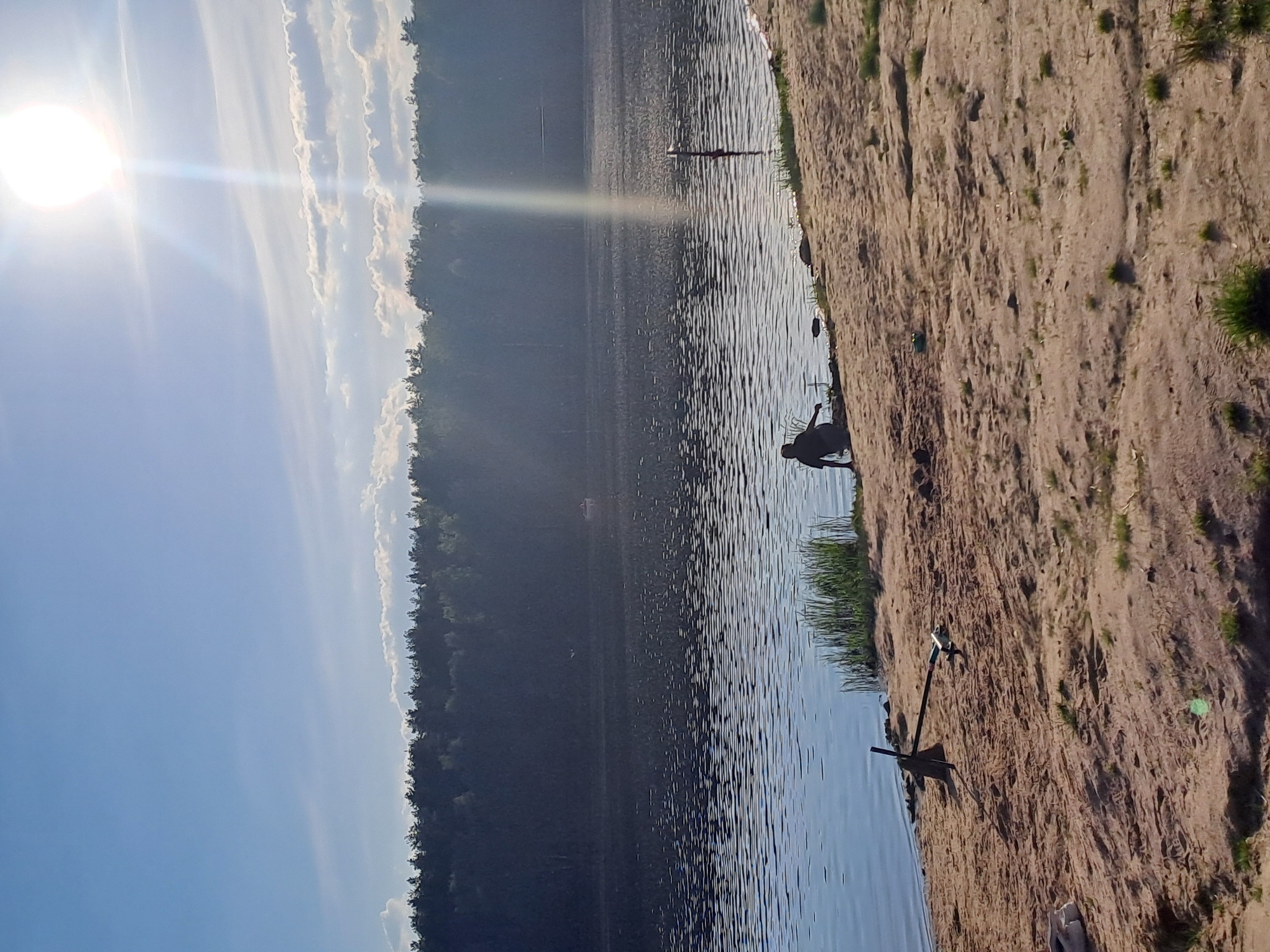 The scene is a bright day at a lake in Меткомелино, Russia. A sandy beach borders the water. A person is standing in the shallow water near the beach. Further out in the lake, a thin post rises from the water. The opposite shore is lined with dense trees. The sky is blue with white clouds and the sun is shining brightly. An anchor is on the beach.