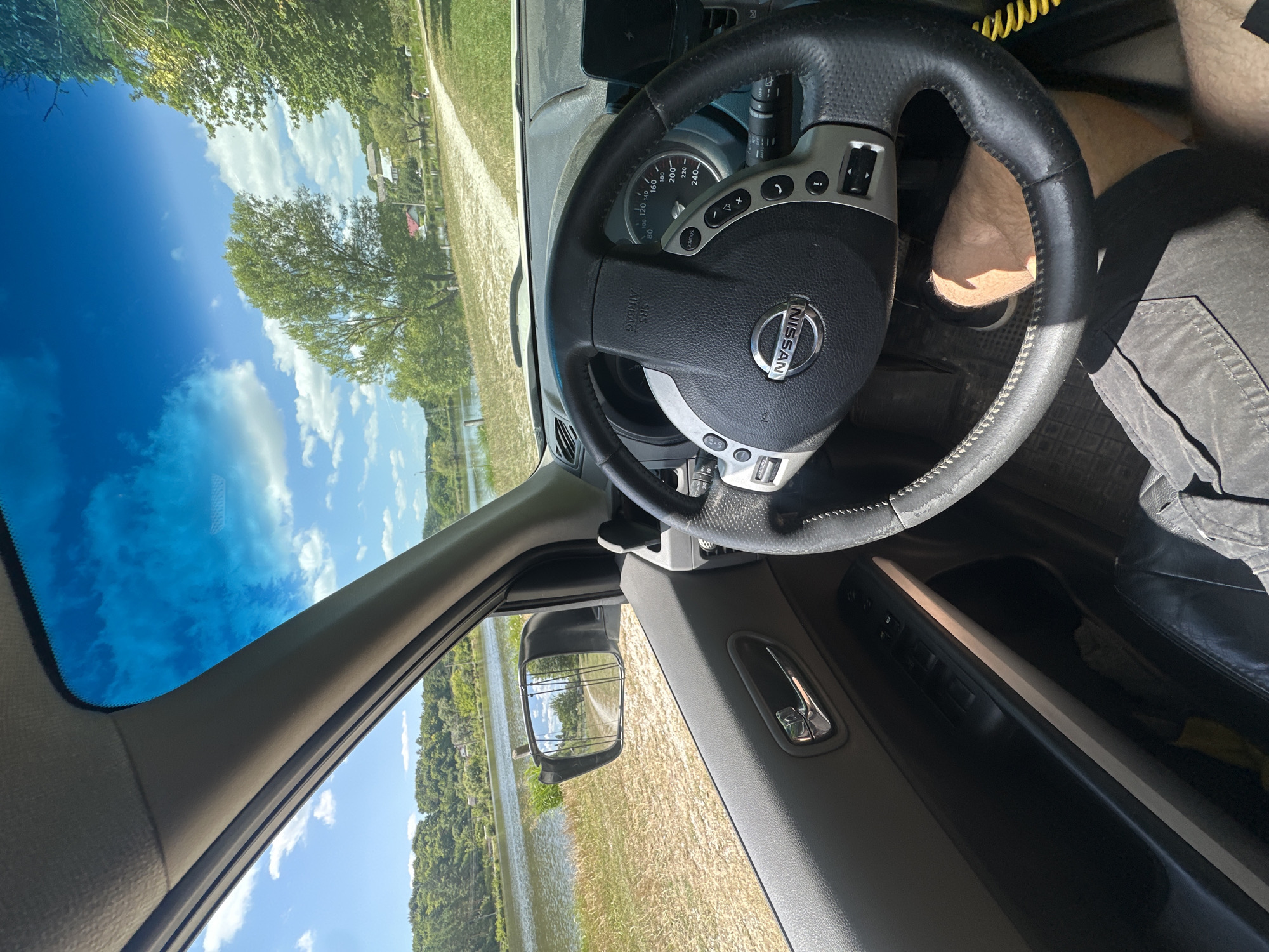 The image is taken from inside a Nissan vehicle, looking out the driver's side window towards a body of water and a tree-lined landscape. The steering wheel and dashboard are visible in the foreground. The scene appears to be a rural area, possibly near the city of Makiv, Ukraine, given the presence of a body of water, trees, and a dirt path. The sky is blue with scattered clouds, indicating a sunny day. There is no visible person in the frame except for a leg and foot visible near the pedals.