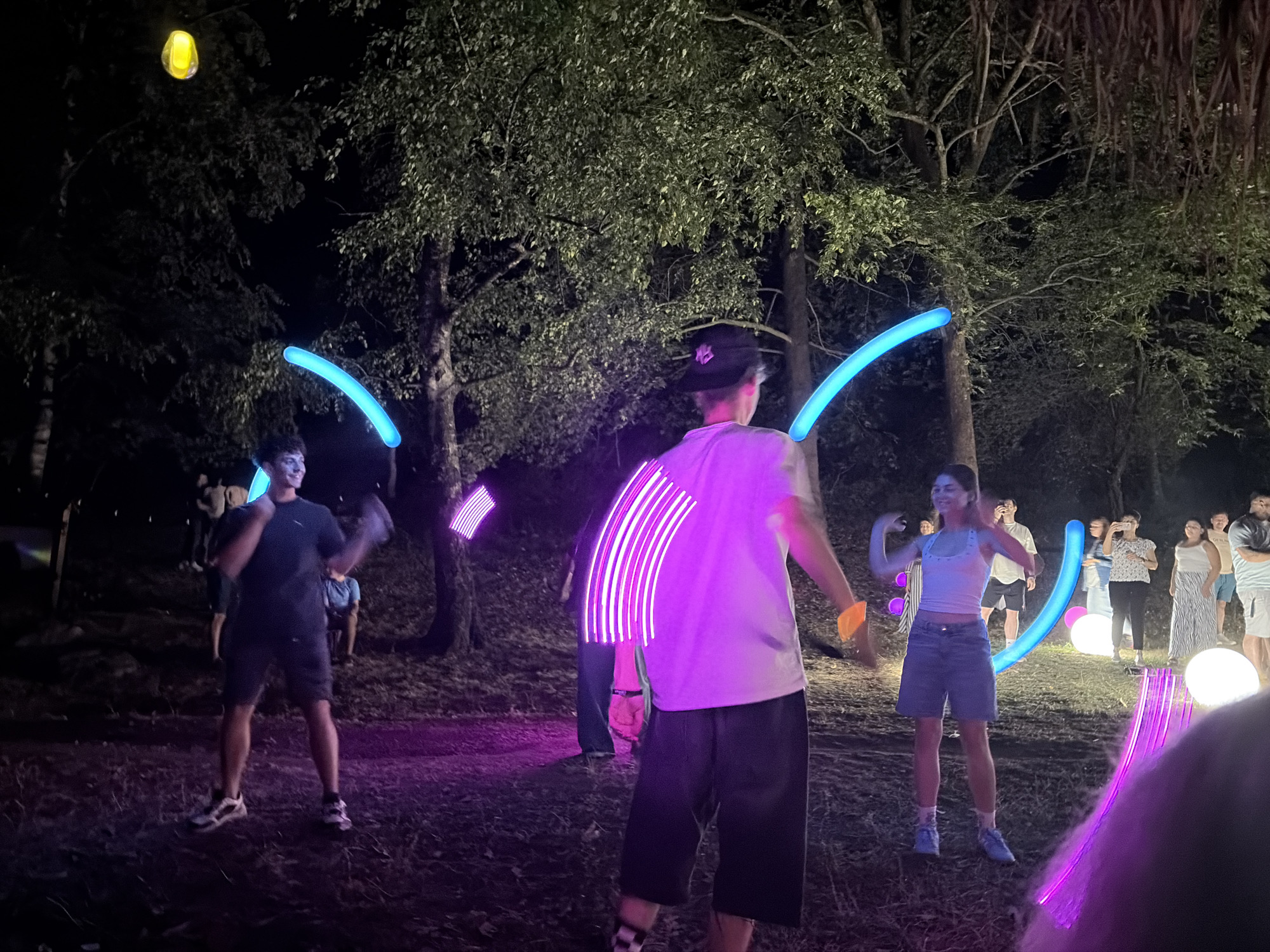 The image depicts an outdoor nighttime event likely taking place in Zgorigrad, Bulgaria, suggested by the provided context. The scene is illuminated by various light sources, including glowing blue tubes held by individuals, purple light effects, and several large white spherical lights on the ground. Trees with dense foliage form a backdrop, creating a somewhat rustic and natural environment.
In the foreground, a person wearing a dark t-shirt and shorts is visible, holding a glowing blue tube. To their right, another individual in a white t-shirt and dark shorts is also interacting with light objects, with purple light streaks emanating from their arms. Further to the right, a young woman in a white top and denim shorts is standing, appearing to be dancing or posing. Several other people are scattered in the background, some holding phones as if taking pictures or videos. The ground is covered with dry leaves and grass. The overall atmosphere appears to be festive and recreational, possibly a festival or gathering.