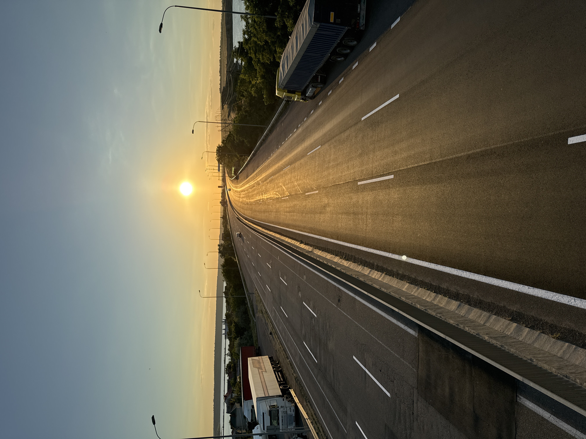 The image shows a highway in Yelyzavetivka, Ukraine, during sunset. The sun is visible in the sky, casting a warm glow on the road and the surrounding landscape. The highway has multiple lanes and is bordered by a concrete barrier and streetlights. Several trucks are parked on the side of the road, and one truck is driving on the highway. In the distance, there is a body of water and some trees. The sky is clear with a gradient of blue and orange colors.