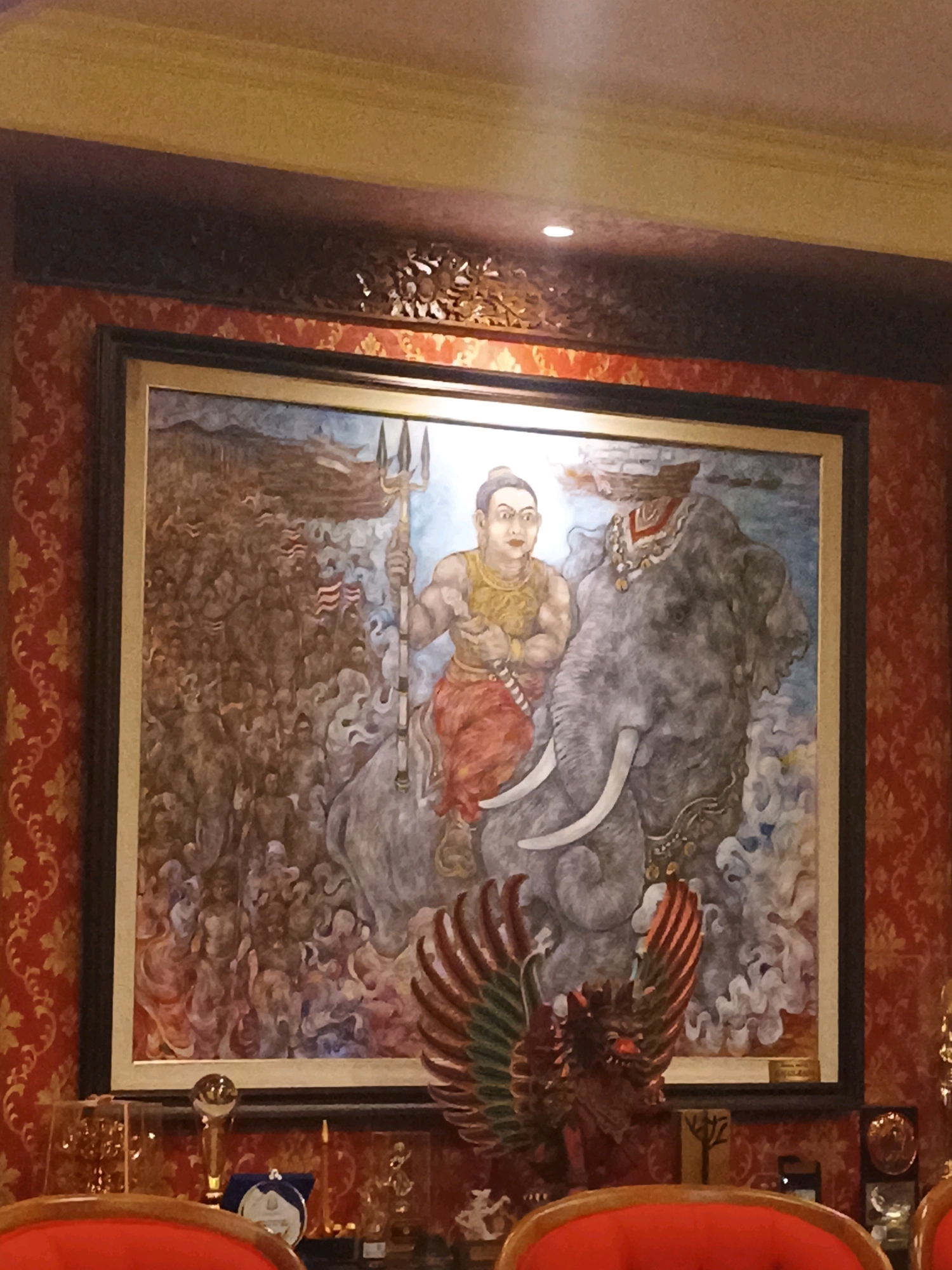 The image displays a detailed, traditional Indonesian mural framed and hung on a wall with red and gold patterned wallpaper. The mural depicts a powerful figure, likely a warrior or deity, riding an elephant. The figure is muscular, adorned in a golden vest and red trousers, and holds a trident. The elephant is large and grey, with prominent tusks and decorated with ornate jewelry. Behind the elephant, a multitude of figures and elements suggest a battle or grand procession, with some figures carrying flags. The mural is illuminated by a spotlight from above. In front of the mural, on a shelf, are various trophies and decorative objects, including a golden chalice and a circular medallion. Red chairs are visible in the foreground, suggesting this is a display area in a public or private establishment. The style and subject matter point towards Balinese art, likely located in Tabanan, Indonesia, a region known for its rich cultural heritage and traditional arts.