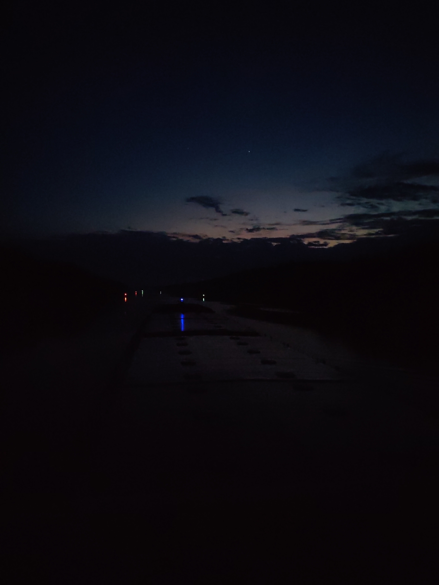 The image depicts a night scene in Aleksandrovskoye, Russia. A road or walkway is visible in the foreground, appearing dark with some lighter-toned patches that suggest concrete or a similar material. There are footprints visible on this surface. In the midground, there are scattered lights, some with a blue hue and others with a reddish hue, likely from streetlights or other artificial sources. The background shows a dark sky with some cloud cover. There is a hint of twilight or the very beginning of sunrise on the horizon, with faint colors peeking through the clouds. Two faint stars are visible in the upper portion of the sky. The overall scene is dim and obscured by darkness.