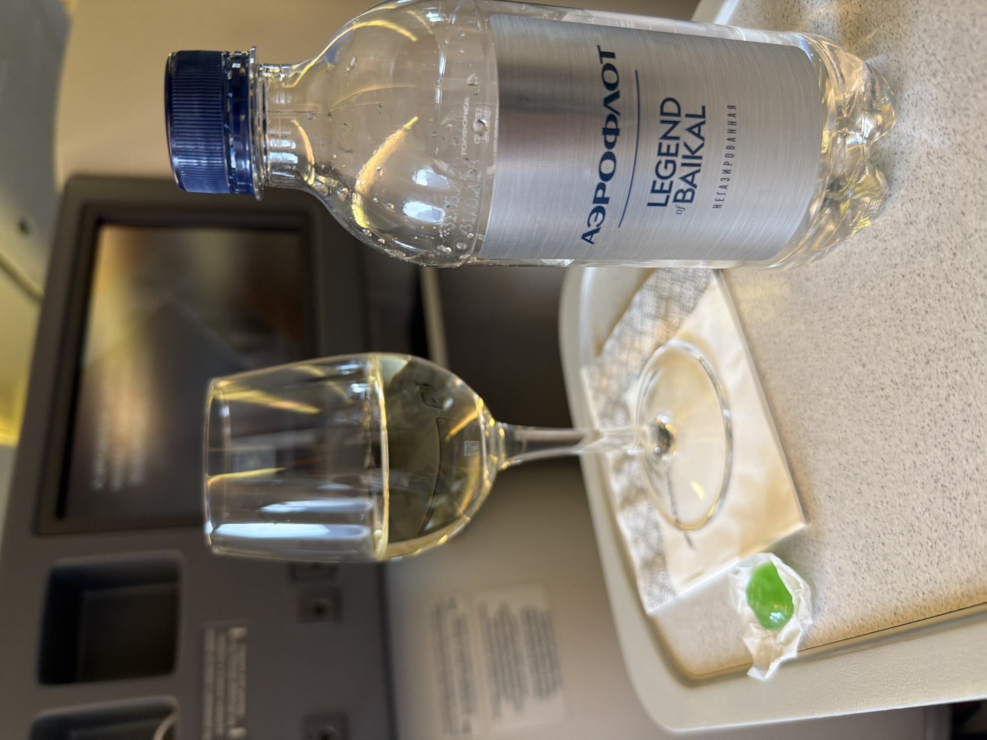 The image depicts a close-up view of a tray table in an airplane cabin, likely during a flight with Aeroflot, as indicated by the branding on the water bottle. On the tray table, there is a stemmed wine glass filled with a pale yellow liquid, presumably white wine. Next to the wine glass is a bottle of "Legend of Baikal" water, which is sealed with a blue cap. The label on the water bottle features the Aeroflot logo. To the left of the wine glass, a small, green, wrapped candy or mint is placed on a white napkin. In the background, the seatback entertainment screen is visible, along with the interior of the airplane cabin, suggesting a travel setting. The overall lighting is dim and warm, typical of cabin lighting. The location is identified as Yelizovo, Russia, which is the site of Yelizovo Airport (ELTZ), a major airport serving the Kamchatka Peninsula. This suggests the photo was taken on a flight departing from or arriving at Yelizovo.