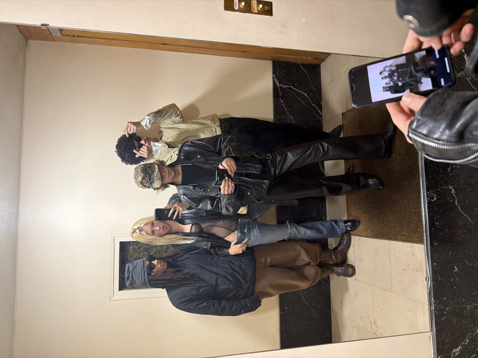 Four people are reflected in a mirror, posing for a photo being taken by someone outside the frame. Starting from the top, the first person has dark curly hair and is wearing a tan jacket. The second wears a camouflage hat and a black leather jacket. The third has blonde hair and wears a sheer top and denim jacket. The bottom person wears a dark beanie and a dark jacket with tan pants. The reflection captures an elevator panel, a patterned floor, and the hand and phone of the person taking the photo.