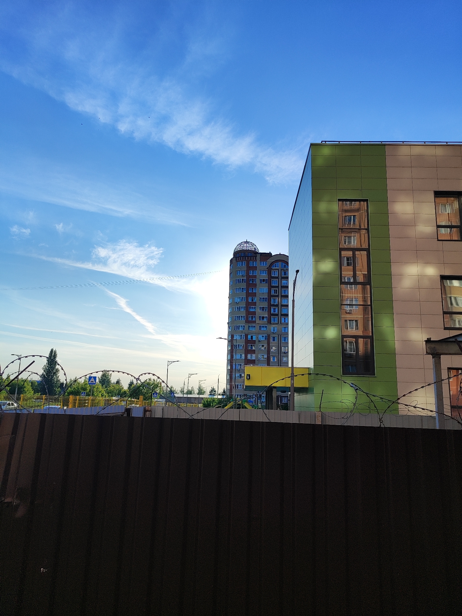 The scene depicts a street in Shchyolkovo, Russia, under a bright blue sky scattered with white cirrus clouds and distinct contrails. The sun, positioned low in the sky, is partially obscured by a tall, cylindrical residential building in the mid-left to background, featuring numerous windows and a prominent domed top. This building exhibits a light blue and reddish-brown facade. To the right, a multi-story building with a contemporary design presents a facade composed of large green and beige panel sections. Its narrow, vertical windows prominently reflect portions of the sky and the cylindrical building.
In the immediate foreground, a dark brown, corrugated metal fence, topped with multiple strands of barbed wire, stretches across the width of the lower section of the image. Behind the fence, an elevated road or bridge structure is visible, characterized by yellow barriers and blue triangular road signs with white pedestrian crossing symbols. Lush green trees and bushes are situated behind this road structure. Several streetlights are interspersed throughout the middle ground. Faintly visible yellow and blue playground equipment is located near the base of the cylindrical residential building. No people are discernible within the frame. The lighting suggests a bright, sunny day, with strong natural light creating reflections and highlights on the architectural surfaces.