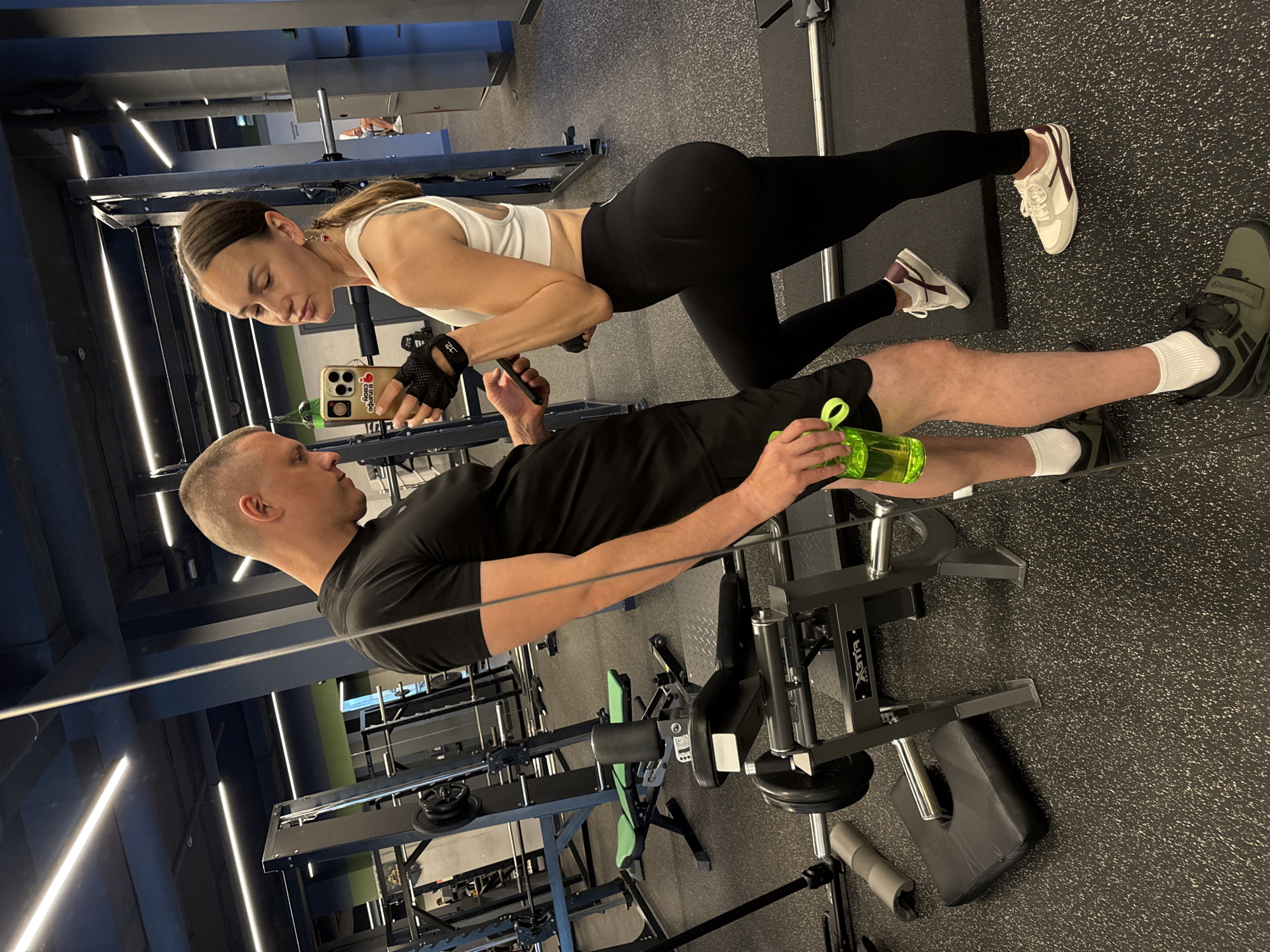 This media file shows two people, a man and a woman, in a gym in Sochi, Russia. The woman is in the foreground, leaning forward with her left leg extended back in a plank position. She is wearing a white sports bra and black leggings, and her hair is pulled back in a ponytail. She is looking at her phone, which she holds in her right hand. The man is behind her, kneeling and looking at the woman. He is wearing a black t-shirt and shorts, and he is holding a green water bottle in his right hand. The gym is well-lit with fluorescent lighting, and the floor is covered in black rubber matting. There are various gym equipment visible in the background, including weight machines and benches. The overall impression is that of a couple working out together at the gym. The lighting suggests it could be daytime or evening. There is no visible weather information. The visible text on the gym equipment is "XFit". The phone in the woman's hand has a case with "Rider" and "Max" written on it, along with some Cyrillic text.
