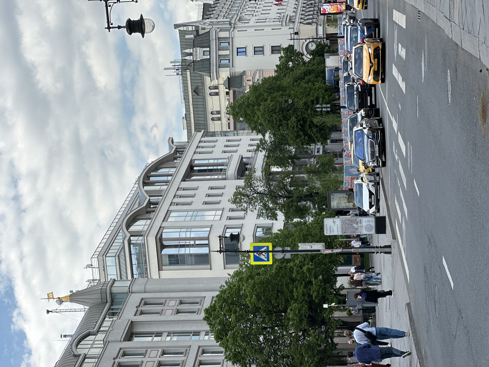 This image shows a street scene in Moscow, Russia. The camera is positioned at ground level, looking upwards along a street lined with buildings on the left and a row of parked cars on the right. The architecture consists of ornate, light-colored buildings. The one closest to the viewer has a spire and gold accents. Another building displays a clock. A crosswalk sign is visible at the street level. Pedestrians are present on the sidewalk, some walking and some standing. The vehicles on the street range from dark-colored SUVs to taxis. The sky is partly cloudy.