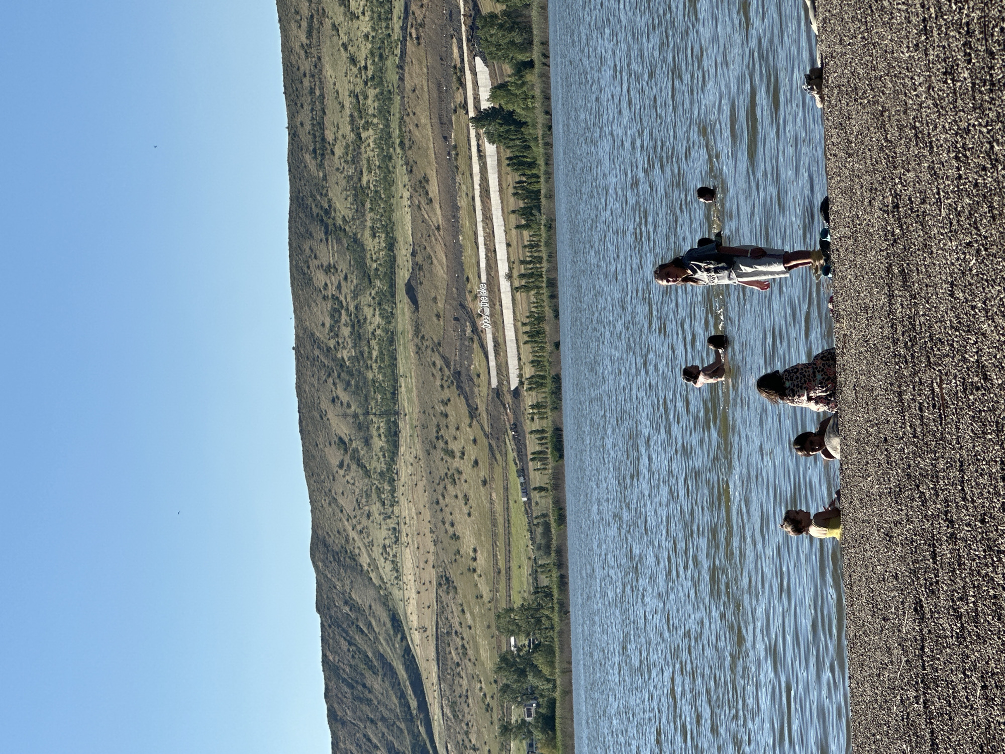 The image shows a group of people gathered by a body of water, with a hilly, grassy landscape in the background. The people are standing near the edge of the water, with some appearing to be in the water. The landscape is characterized by rolling hills covered in dry grass and sparse vegetation. There are some trees and a road visible in the middle ground. The sky is clear and blue, suggesting a sunny day. The overall impression is a casual outdoor gathering in a natural setting.
The image is oriented vertically, with the water and the people on the right side, and the landscape on the left. A small dark object, possibly a bird, is visible in the sky. The people appear to be engaged in conversation or enjoying the scenery. One person in the water is wearing shorts and a t-shirt. Another person is wearing a yellow shirt. The ground in the foreground is covered in small pebbles or gravel. There is some text visible on a road or path in the background, which appears to read "© thelake".
The scene is set outdoors, likely during daytime due to the bright sunlight and clear sky. The location is identified as Tbilisi, Georgia. The mood appears relaxed and informal.
