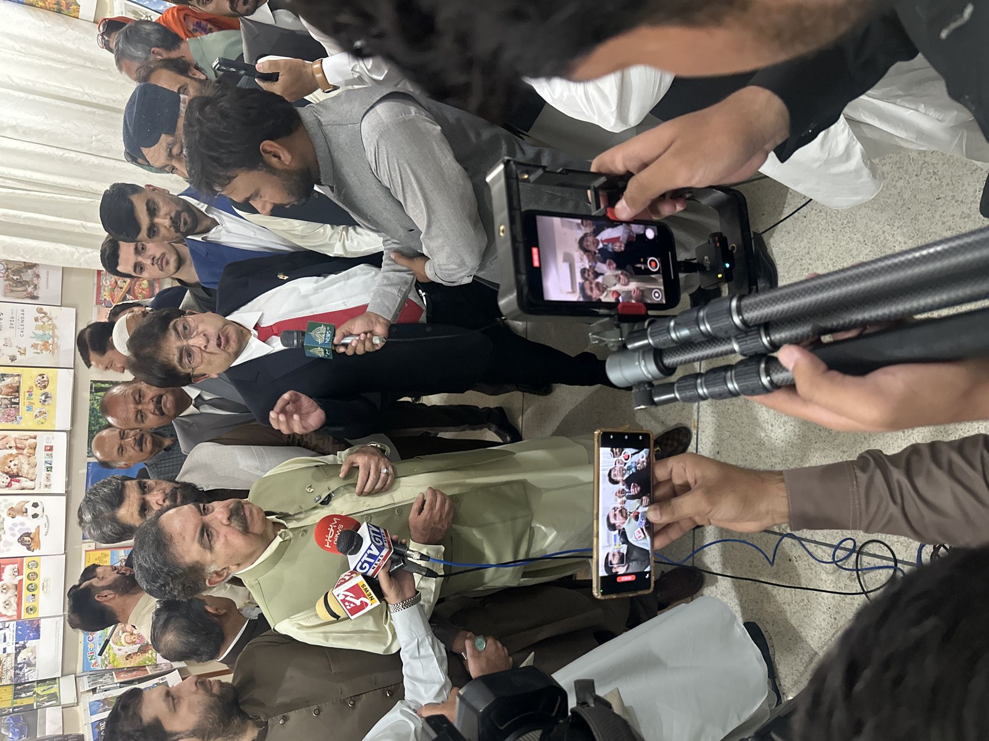 The image shows a group of men, likely politicians and journalists, in what appears to be a press conference. The location is possibly within Quetta City Tehsil, Pakistan, although this is not explicitly confirmed by the visual content. Two central figures are being interviewed, one wearing a light green shalwar kameez and the other a suit with a red tie. Multiple microphones from various news outlets are positioned in front of them. Several people are holding up mobile phones, either recording the event or broadcasting it live. The background includes bookshelves with children's books. The floor is light-colored and possibly tiled. Electrical wires from equipment are visible on the floor.
