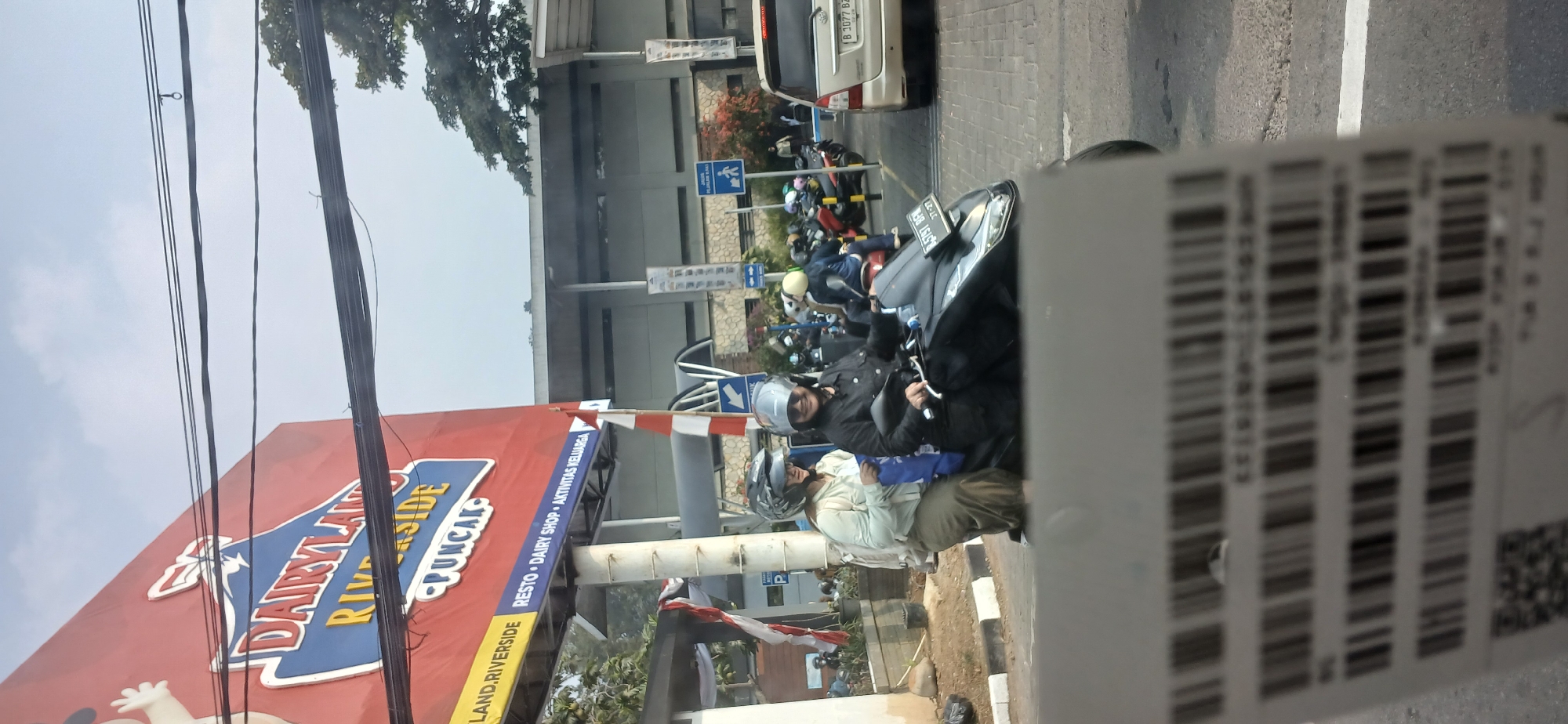 The image, taken in Megamendung, Indonesia, shows a street scene with a prominent "DAIRYLAND RIVERSIDE PUNCAK" billboard in the background. Two individuals are riding on a scooter, both wearing helmets. The person in front, wearing a black jacket, appears to be smiling and driving. The passenger behind them, in a light green jacket, is looking forward. In the middle ground, there are several other scooters and a gold-colored SUV. The environment appears to be a typical urban or semi-urban setting with buildings and trees visible. A blurred foreground element suggests the photo may have been taken from inside a vehicle.