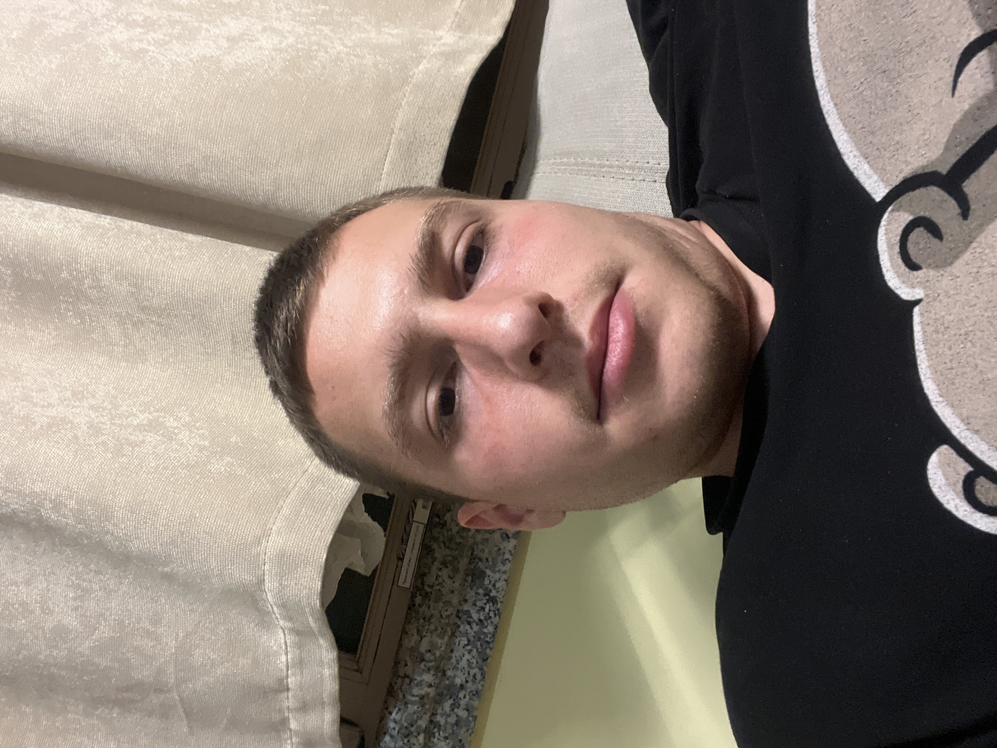 A light-skinned man with short brown hair and stubble is lying down. He's wearing a black shirt with a white cartoon image on the right side of the frame. His face is centered, and he is looking directly at the camera. Behind him, on the left, is a light-colored curtain. Underneath the curtain, there appears to be a marble-like surface next to a doorframe. The location is unknown and could be in Burgas, Bulgaria, or anywhere else.